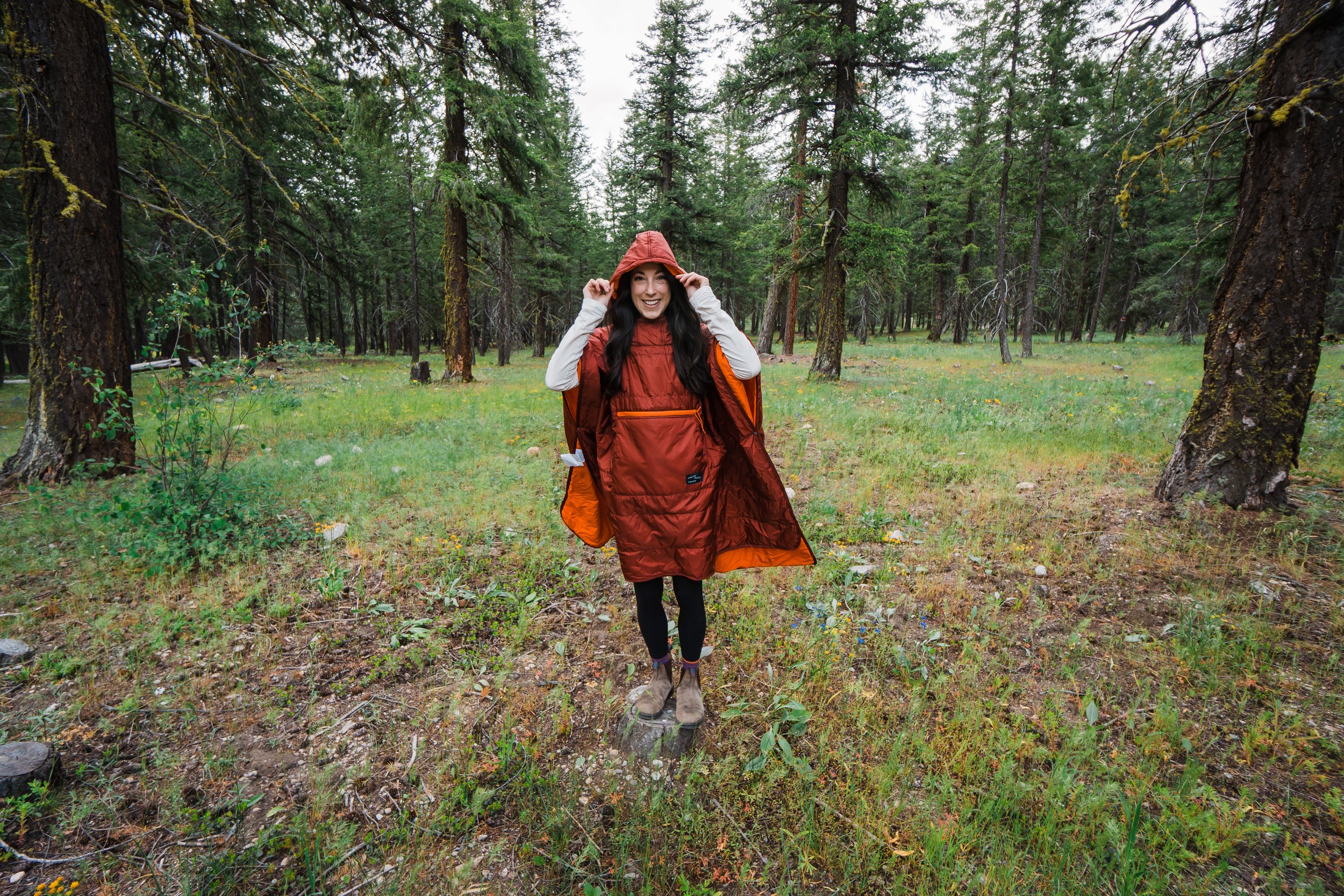 A woman in a red raincoat and hiking boots smiling in a forest with tall trees and green grass.