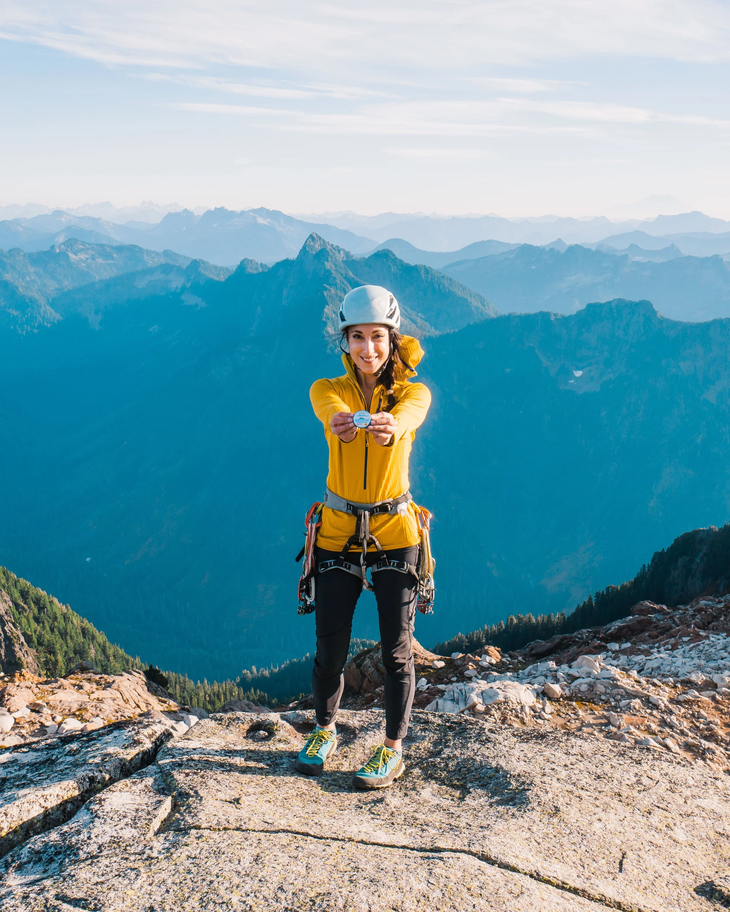 A woman in a yellow jacket and climbing gear standing on a rocky mountain peak, smiling and holding a compass with mountain ranges in the background.