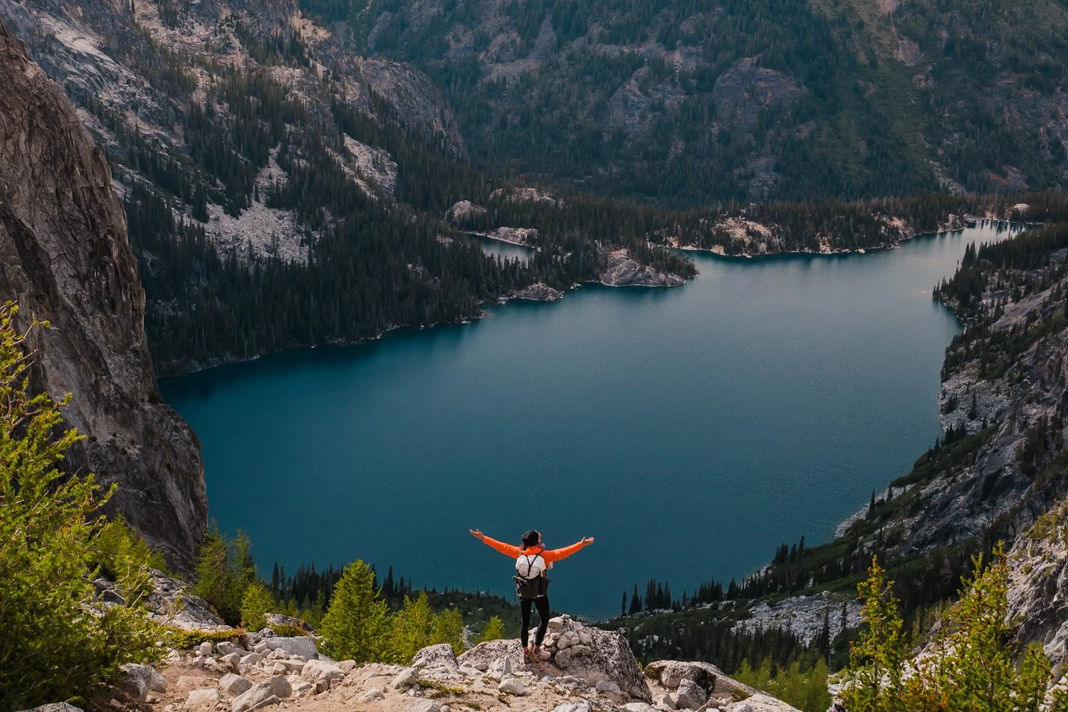 a hiker on aasgard pass in the enchantments with her arms outstretched and a blue cokchuck lake below