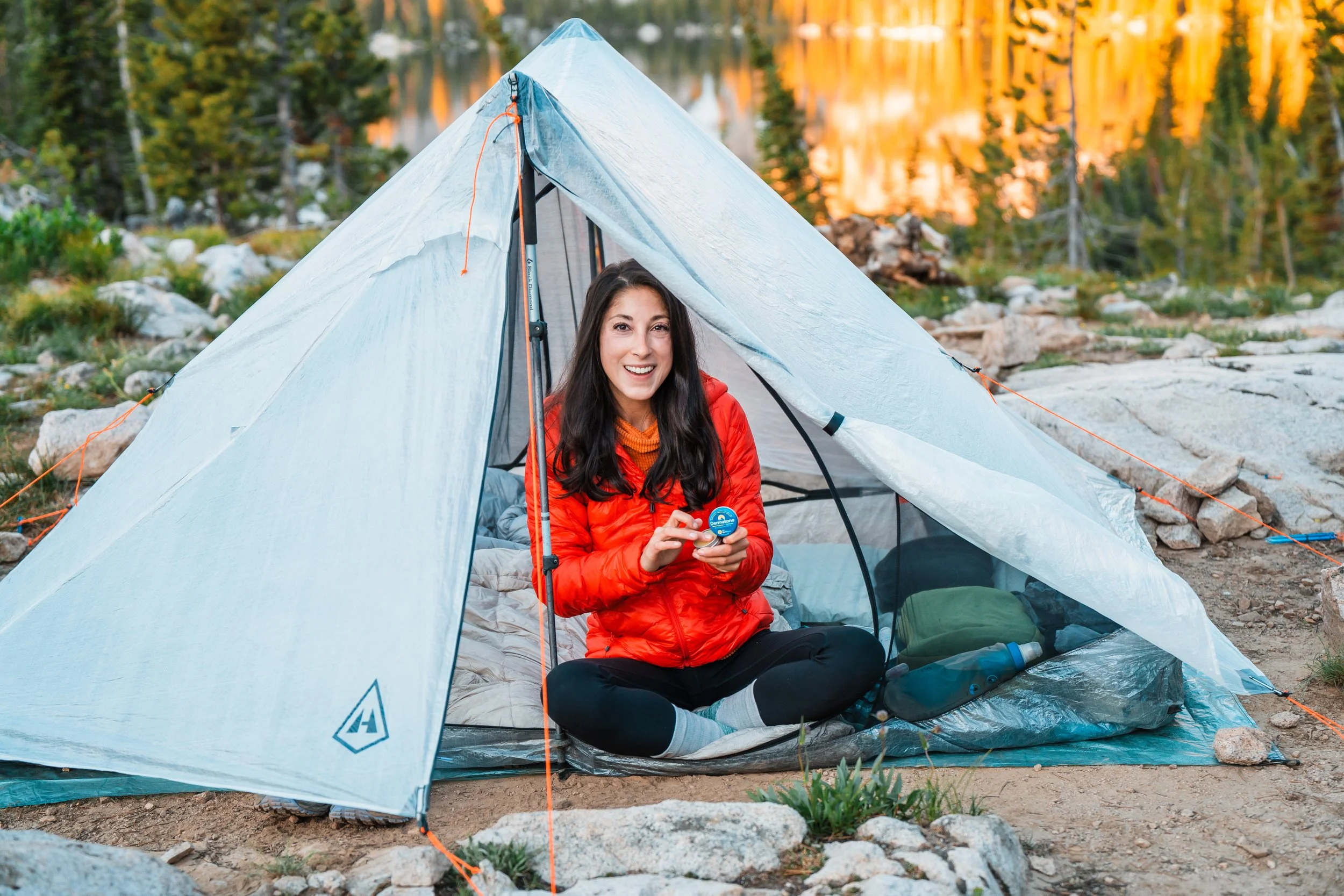 Woman sitting inside a tent camping in a forest, holding a small container, with a smile on her face, surrounded by rocks and trees, with a lake in the background.