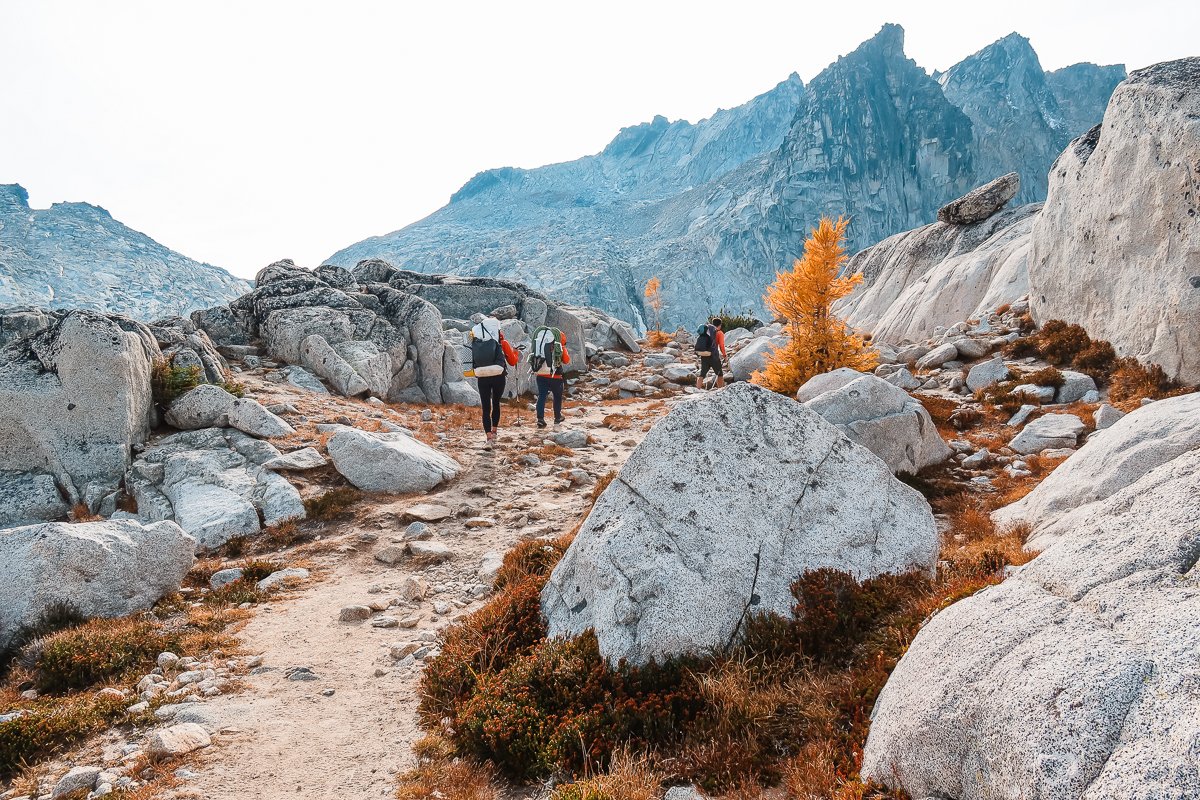 backpackers hiking on a rocky trail, surrounded by boulders and with rocky peaks in the distance in the core enchantments