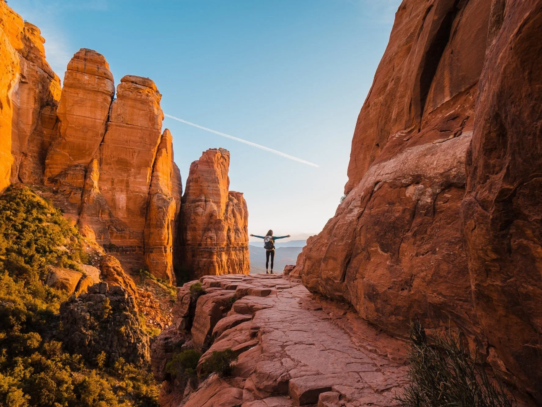 a hiker standing on the edge of a rock cliff with her arms outstretched. Red sandstone rocks on either side.