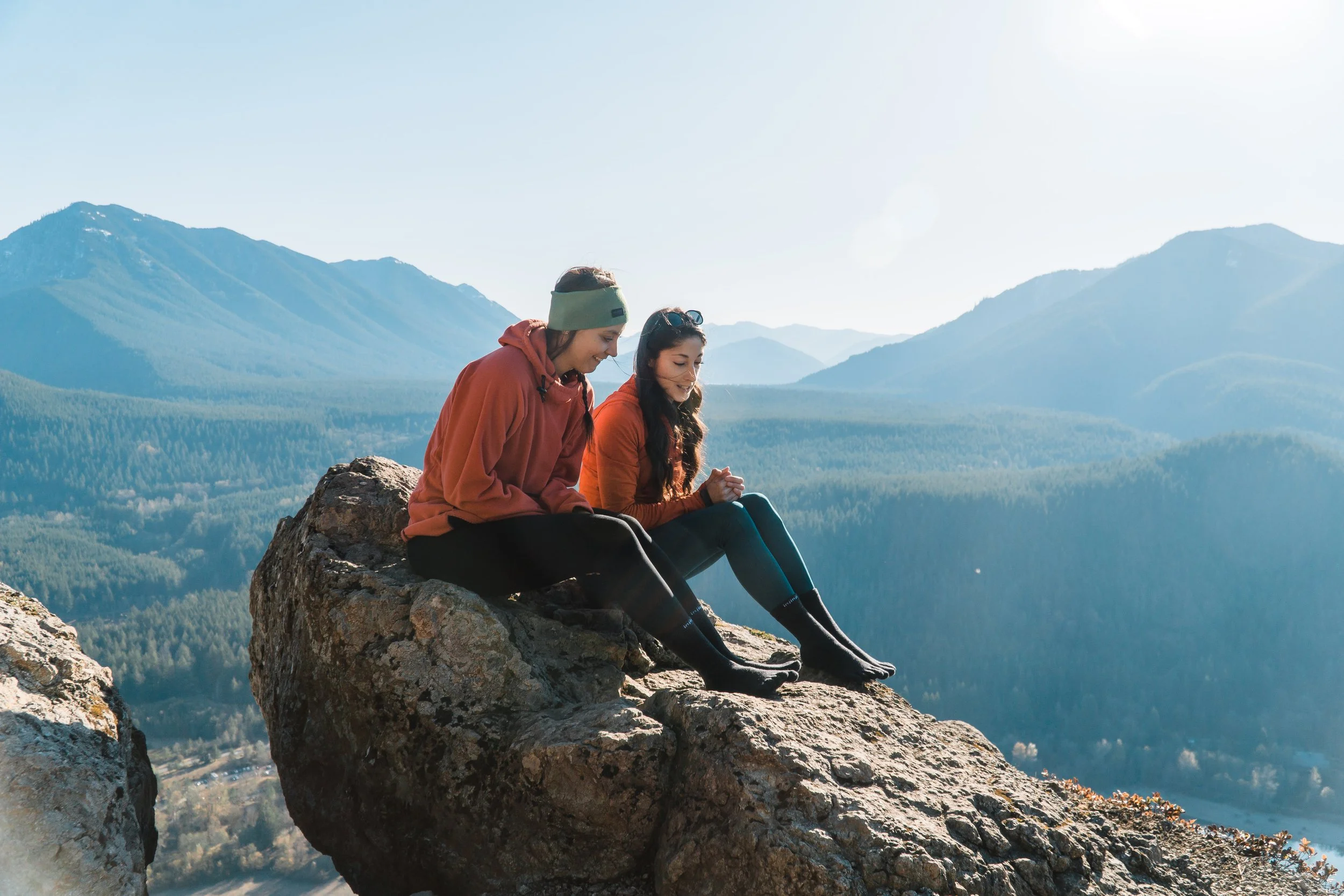 Two women sitting on a large rock overlooking a mountain range with forests, smiling and enjoying the scenery on a clear day.