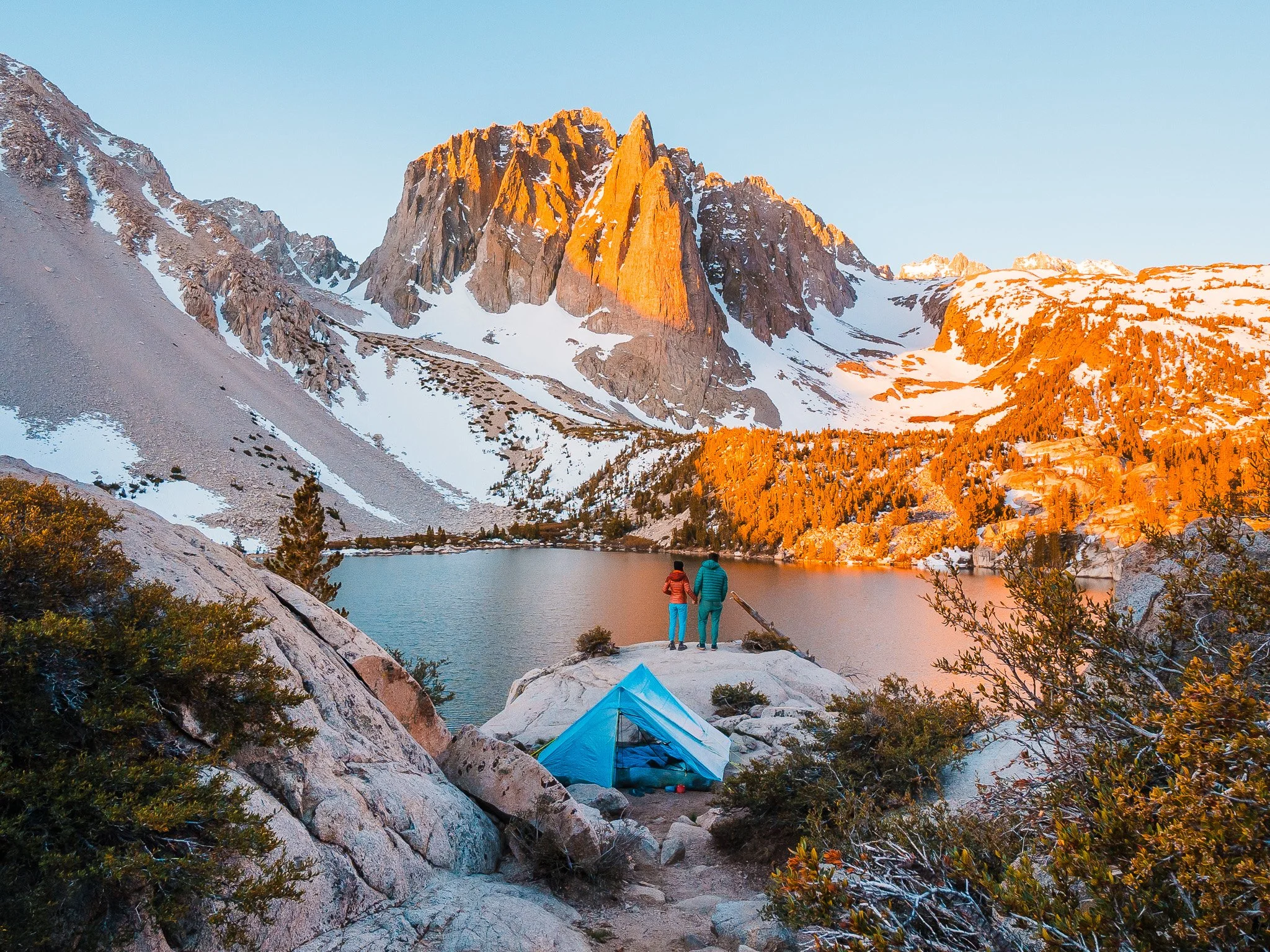 a blue tent and a couple standing behind it on a rock, overlooking a blue alpine lake and the sunrise glow on a large rocky peak in Big Pine Lakes, California