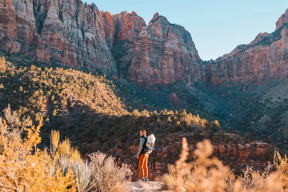 A woman standing at a view point on the watchman trail in zion, with a white backpack and red rocks in the background