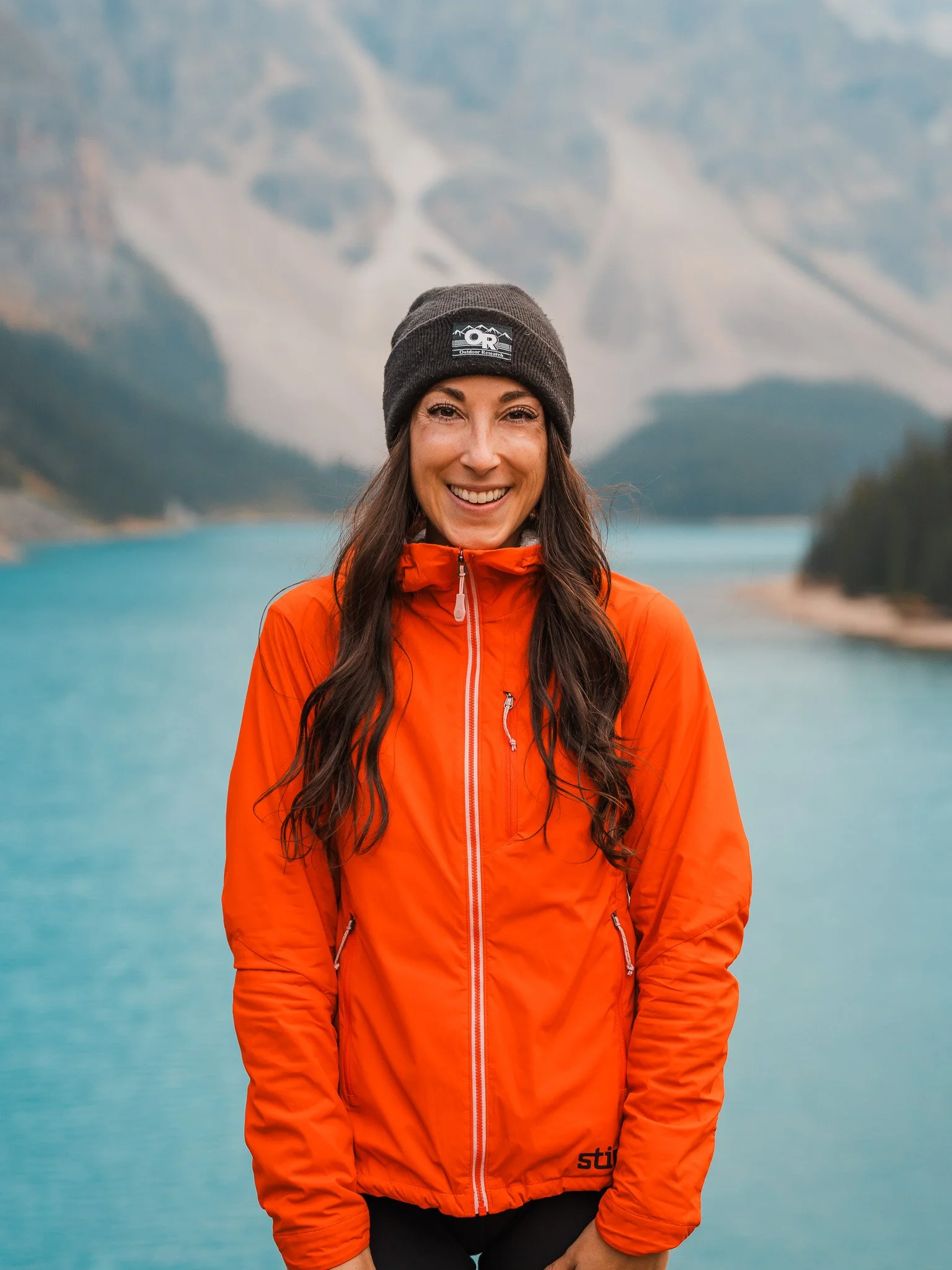 a women in a red jacket and black beanie with long brown hair, smiling and standing in front of Moraine lake in Banff