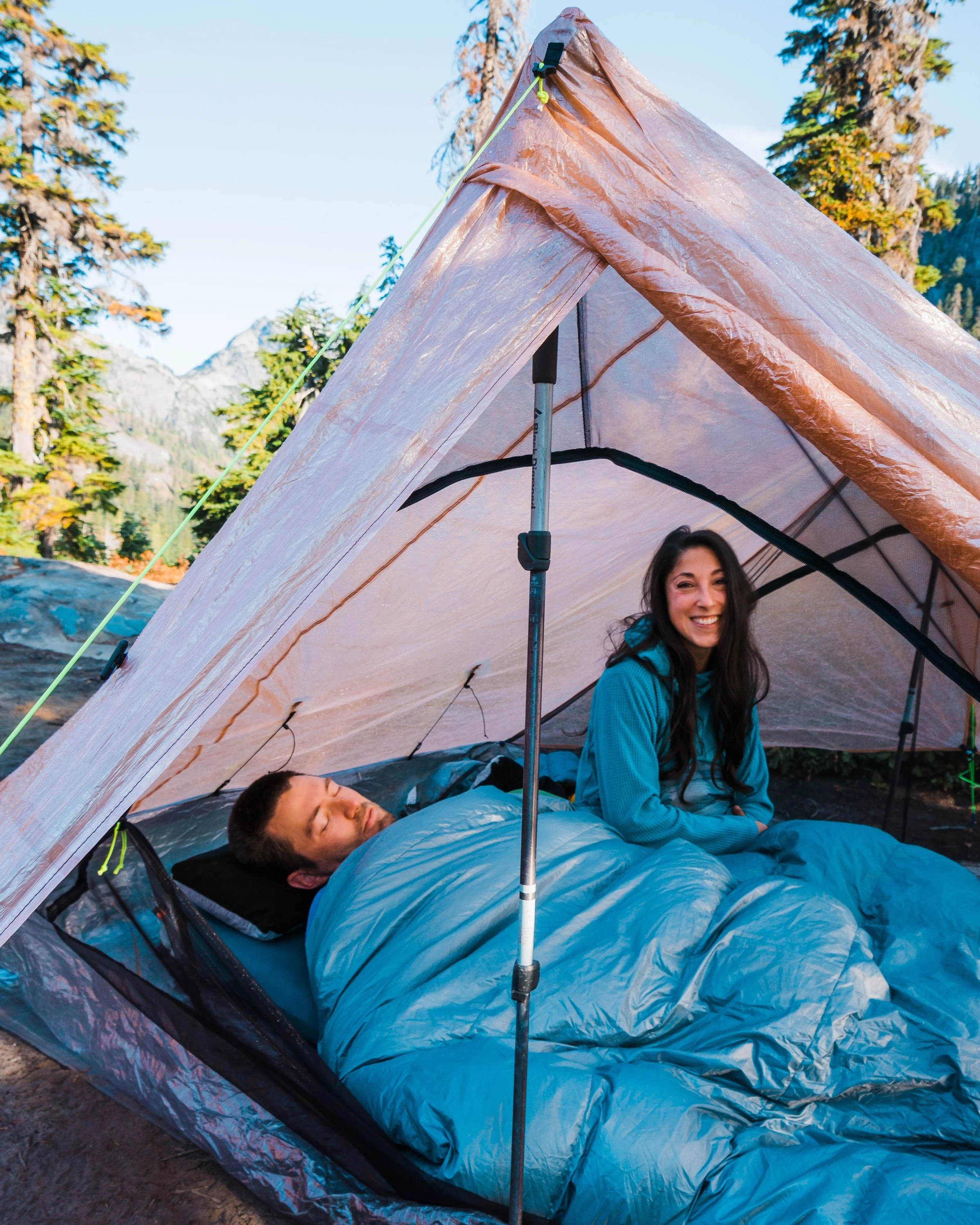 A couple camping outdoors under a tent with trees and mountains in the background. One person is sleeping inside the tent, while the other is sitting up and smiling.