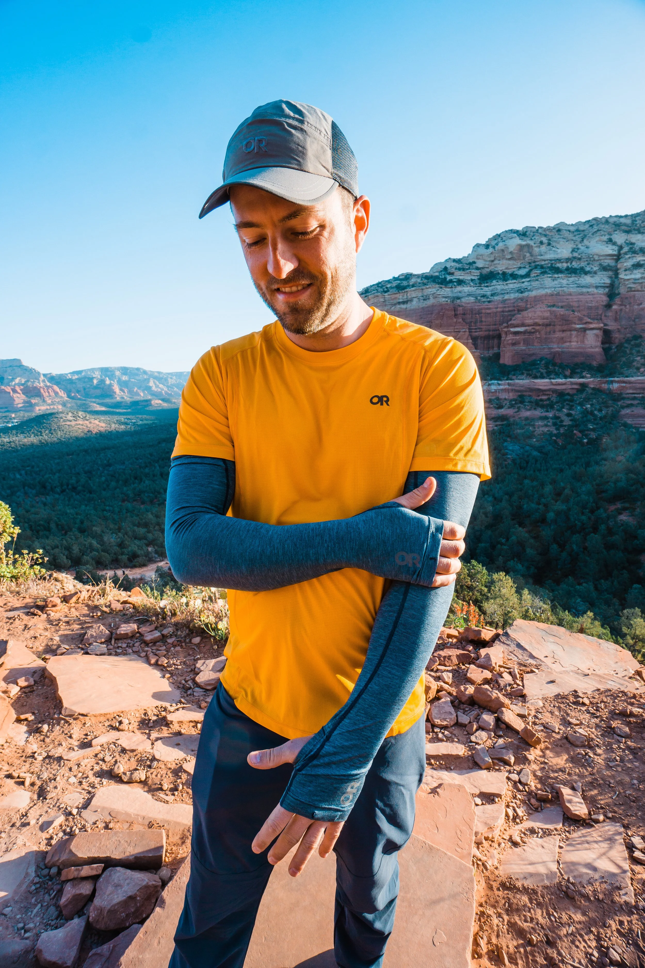 A man in a yellow shirt and gray cap standing outdoors in a desert landscape with rocky terrain, mountains, and clear blue sky, adjusting his blue sleeves.