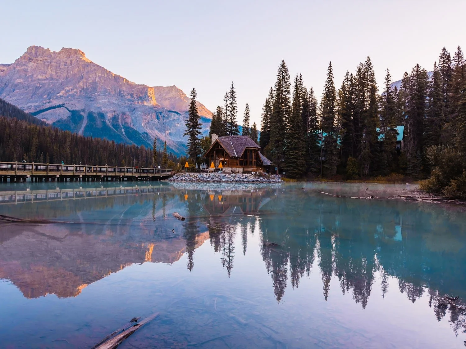 A tiny cabin like building across a bright blue Emerald Lake, with mountains in the background turning pink and purple as the sun rises