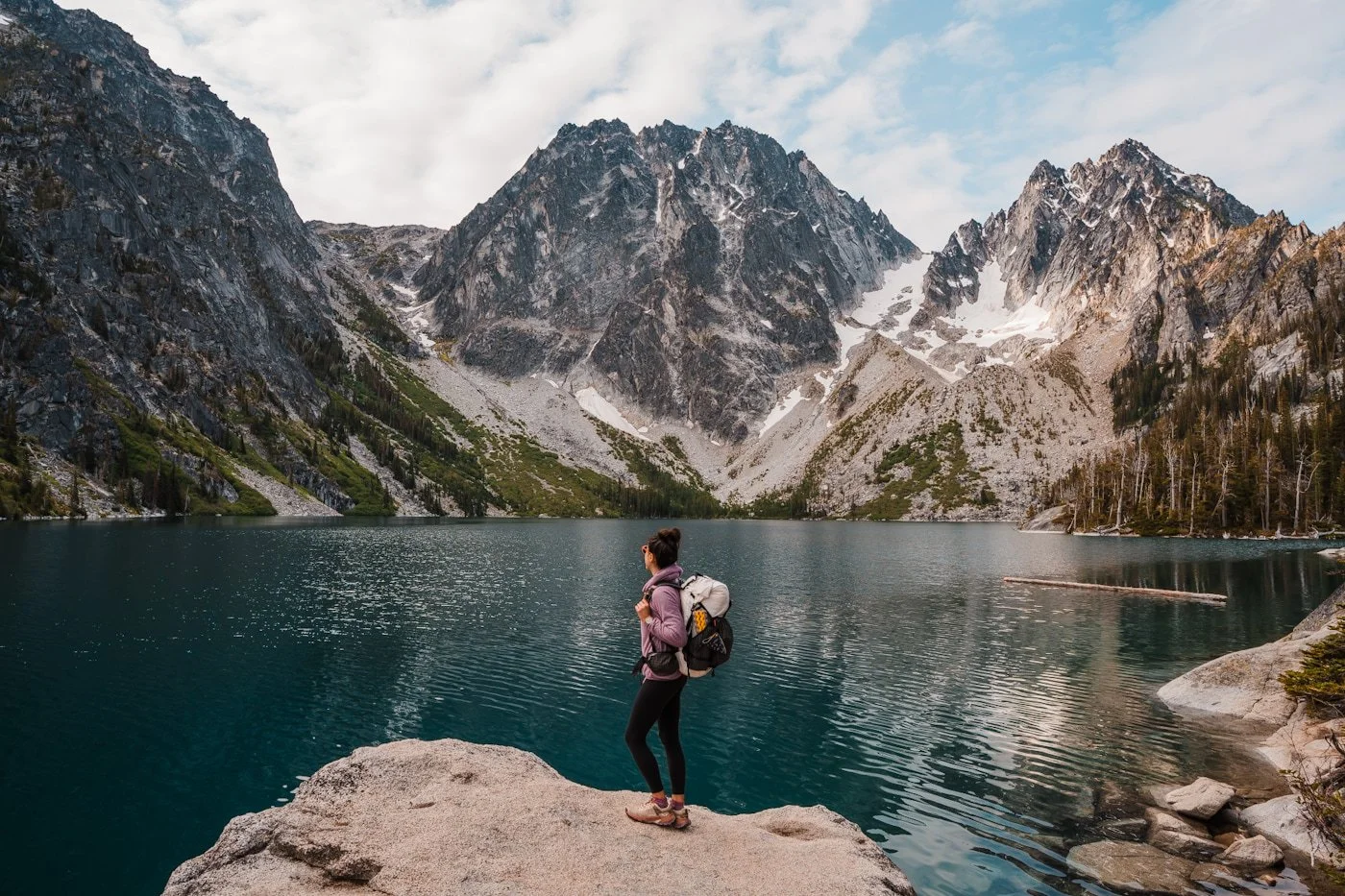 a woman backpacker standing on a rock in front of colchuck lake, a blue alpine lake with rocky peaks in the background
