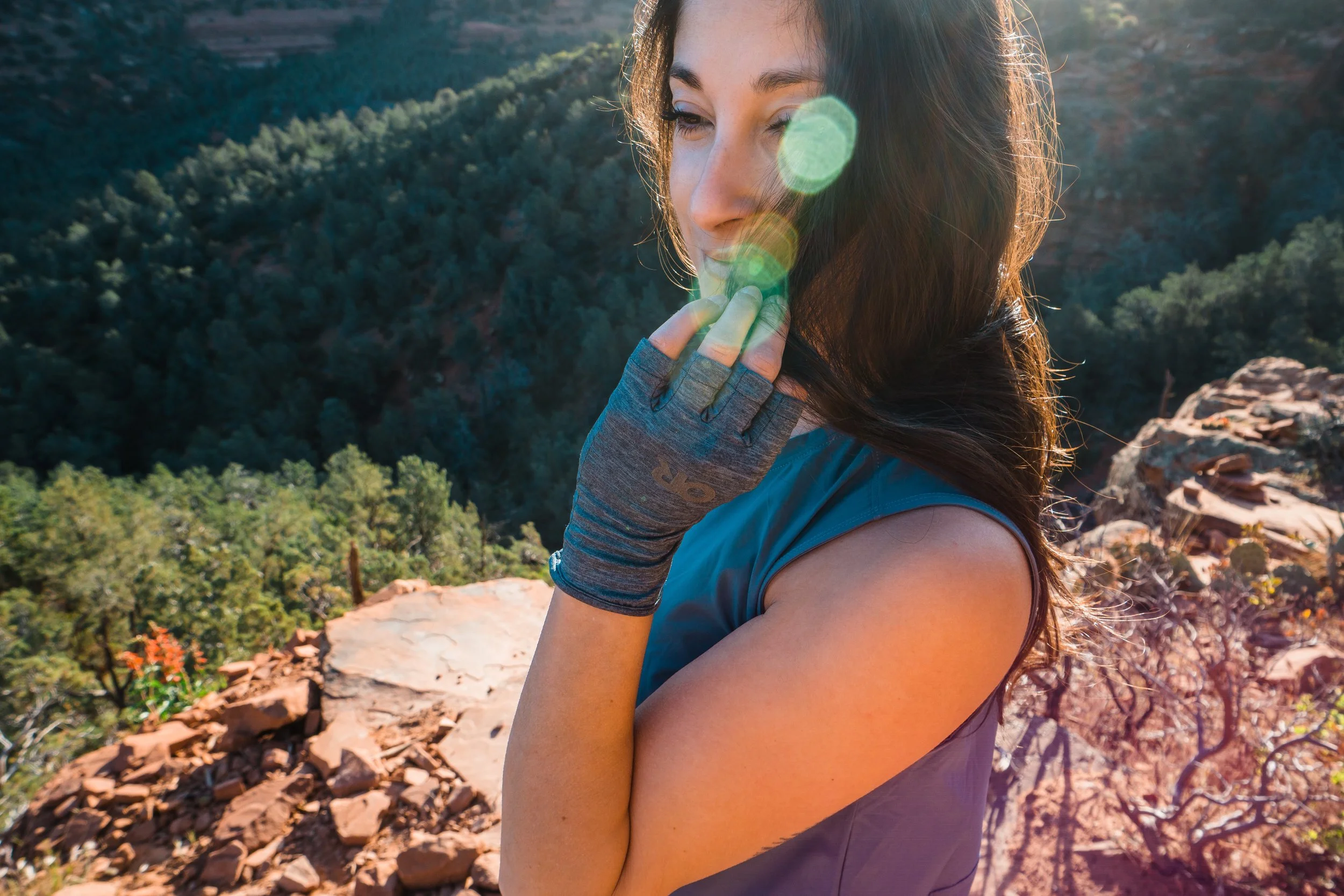 A woman with long dark hair wearing a blue sleeveless top and fingerless gloves is standing on a rocky ledge overlooking a forested canyon during sunrise or sunset.