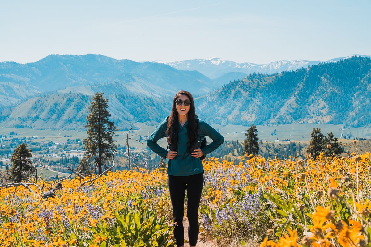 a woman standing on a hiking trail, smiling at the camera, and surrounded by yellow balsamroot and purple lupine wildflowers in Cashmere, Washington