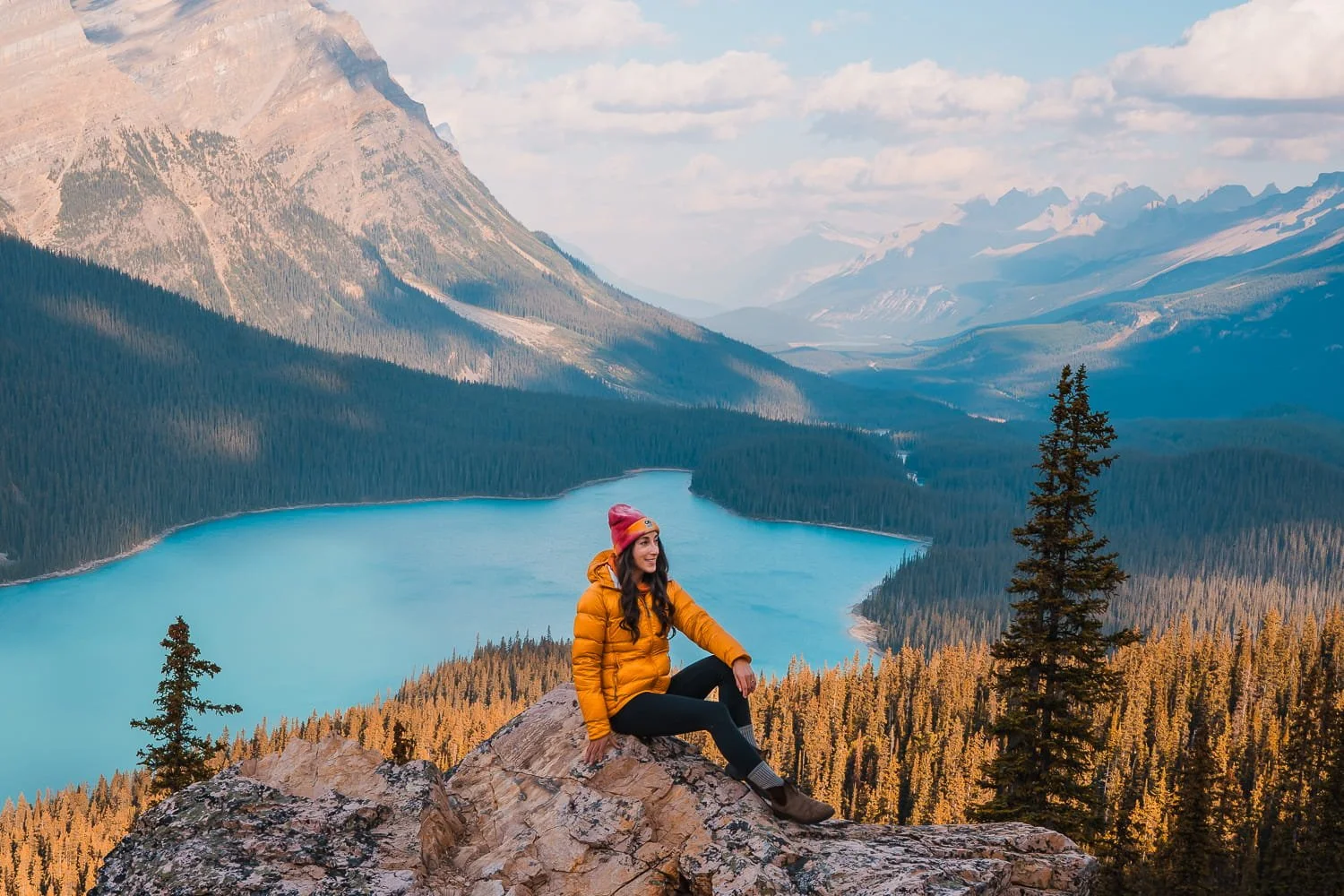 a woman sitting on a rock above Peyto Lake in Banff National Park, wearing a yellow down puffy and pink beanie, with the bright blue lake and mountains in the background