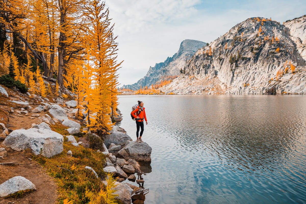 a backpacker in a red jacket and black and white pack, standing on a rock overlooking one of the enchantment lakes with golden larches