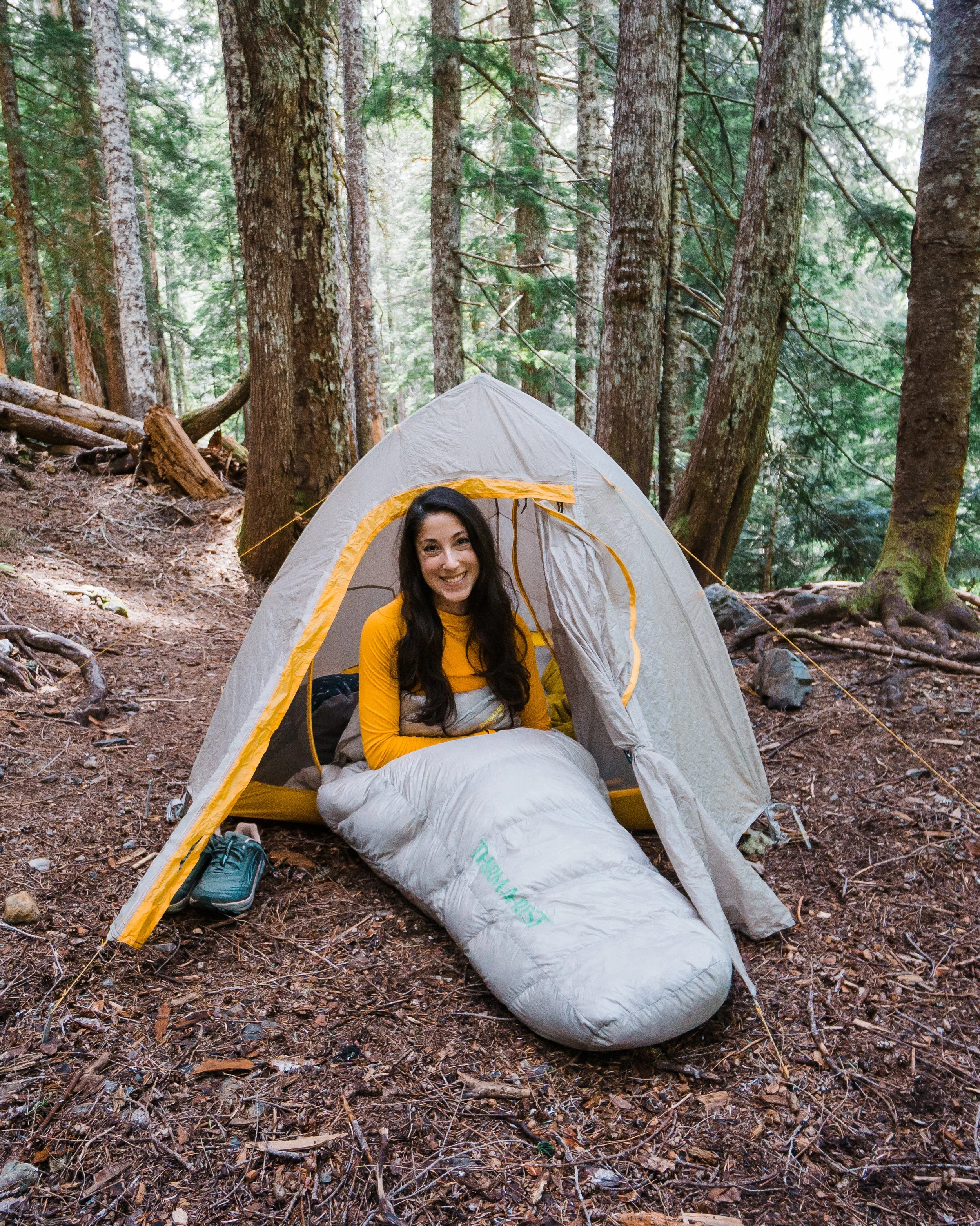 A woman smiling inside a tent in a forest, with trees surrounding her and her sleeping bag in front.