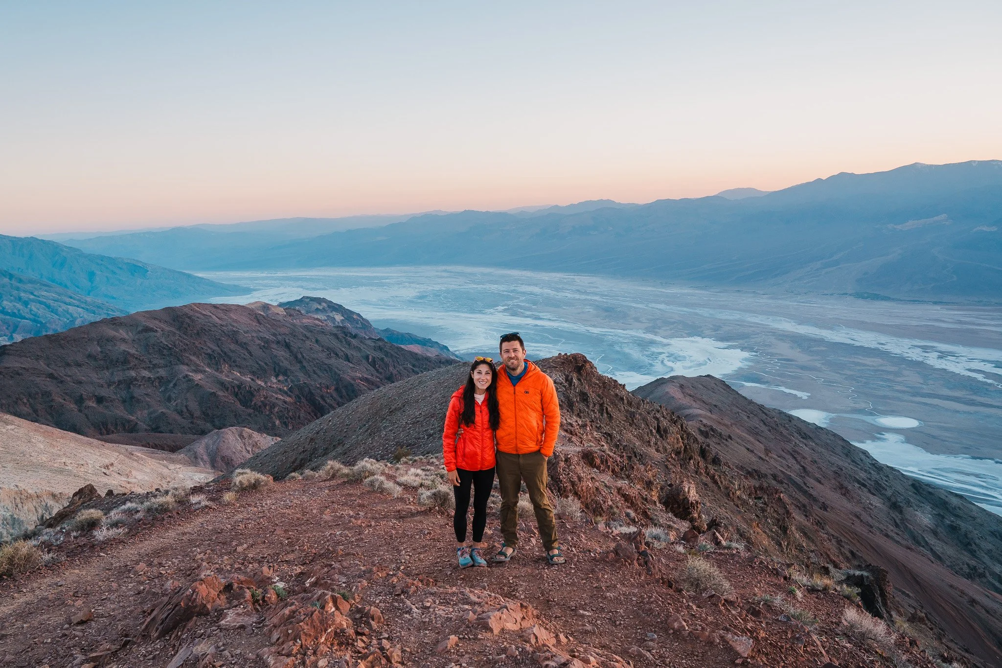 a couple in bright jackets standing at a desert view point at sunset, overlooking the valley and salt flats below, with mountains in the distance