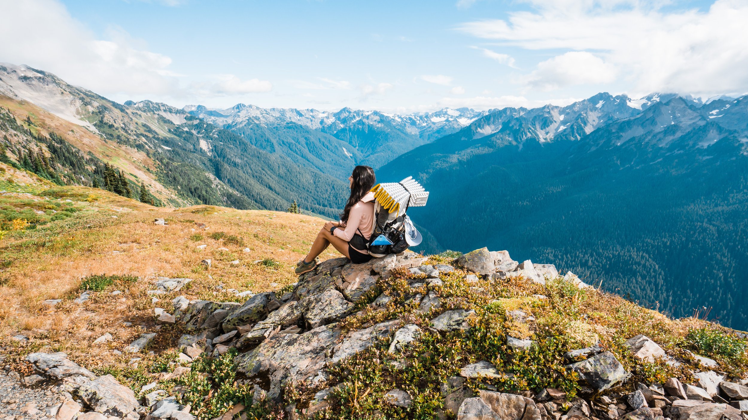A woman sitting on rocks on a mountain, overlooking a vast mountain range with snow-capped peaks, green forests, and a partly cloudy sky.