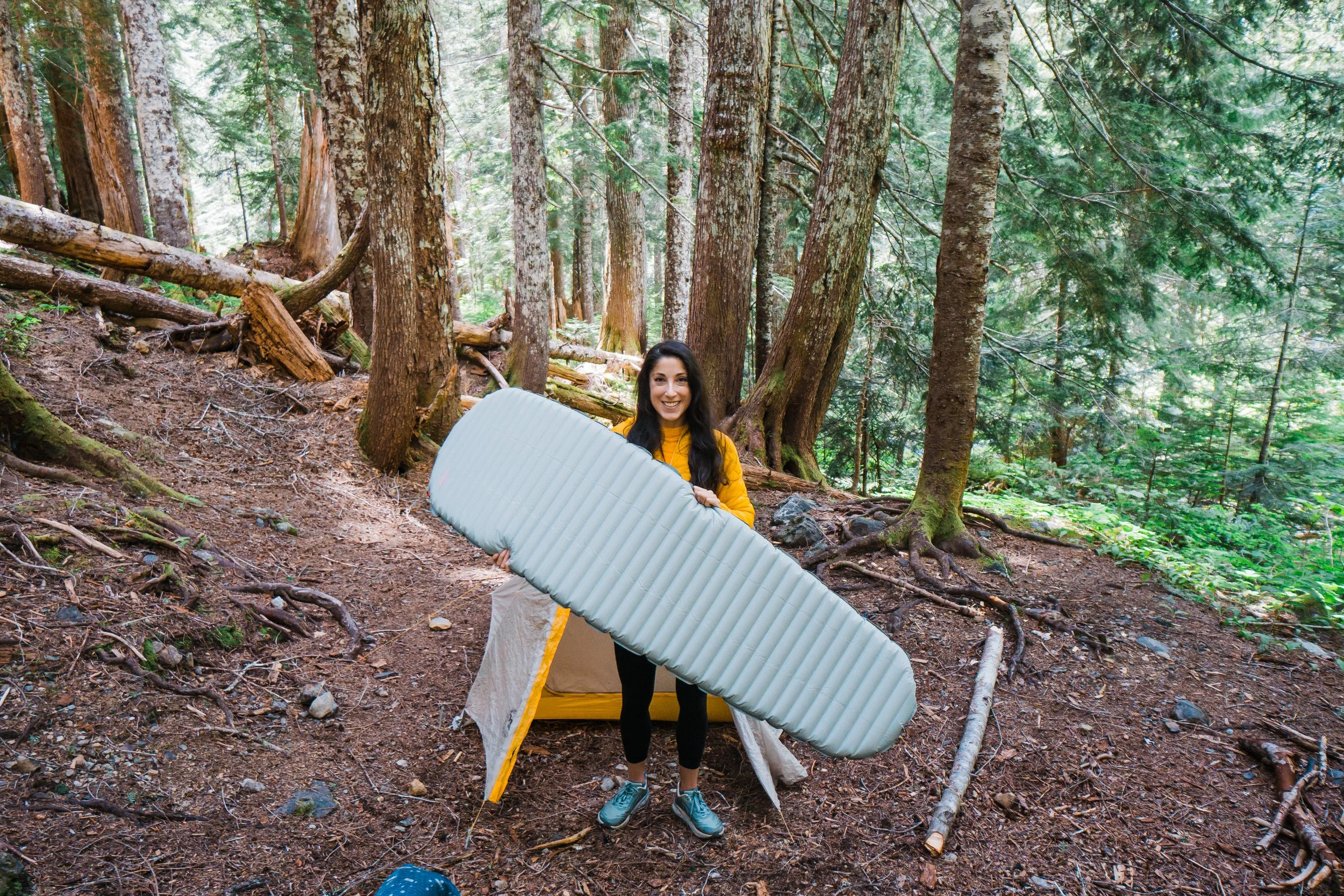 A woman in a yellow jacket holding a gray sleeping pad inside a yellow tent in a forest with tall trees and green foliage.