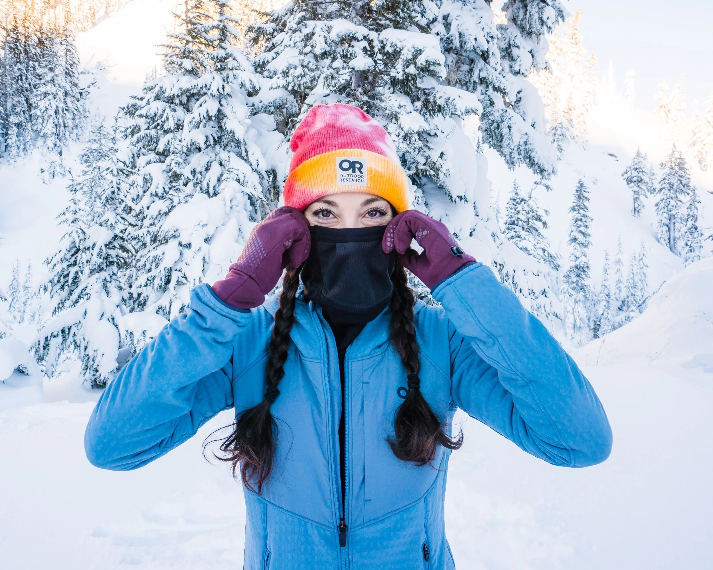 A woman with braids wearing a pink and yellow beanie, black face mask, purple gloves, and a blue winter jacket standing outdoors in a snowy forest.