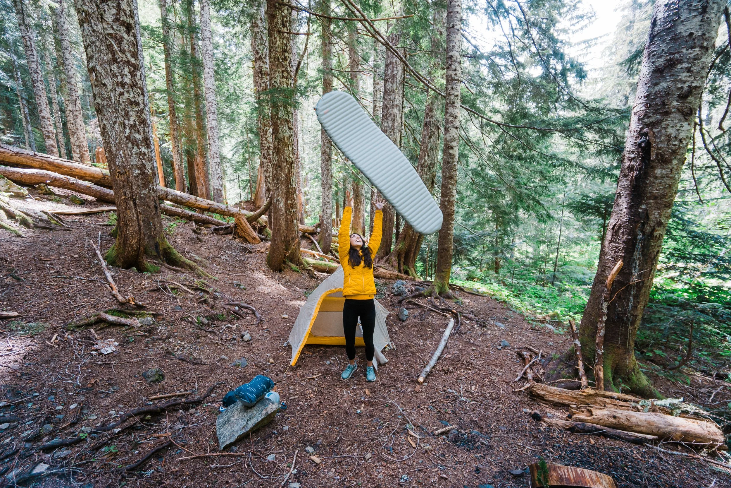 A woman standing outside a camping tent in a dense forest, tossing a large white foam camping mattress into the air. The tent is set up on a dirt ground surrounded by tall trees.
