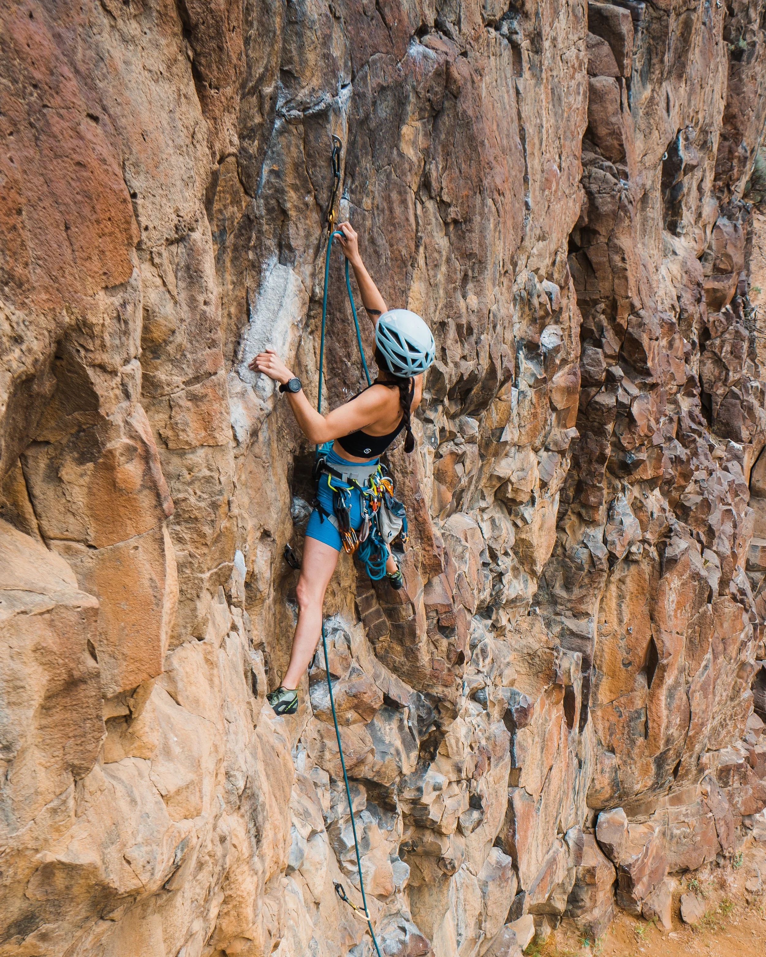 A woman rock climbing on a steep, rocky cliff, wearing a helmet, harness, and climbing shoes, gripping the rock with her hands and feet.