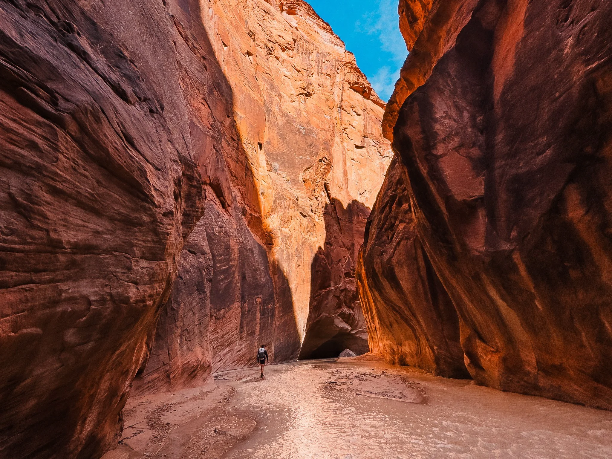 a hiker walking through water in an open section of paria canyon, with towering sandstone walls and sun coming through