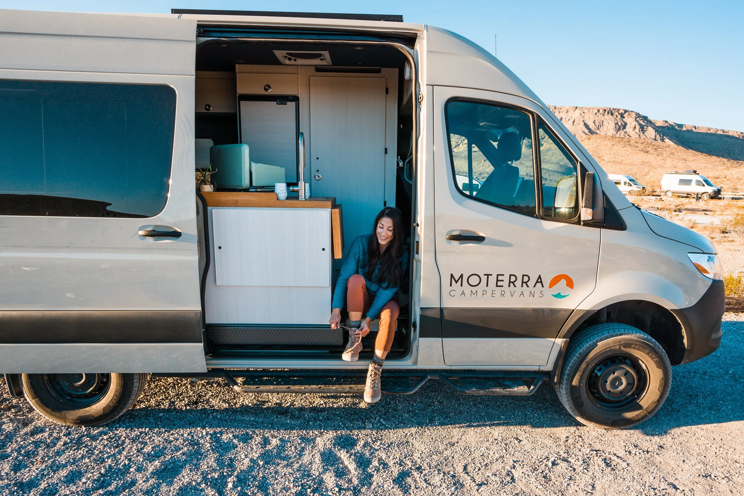 Woman sitting on the steps of a camper van in a desert landscape, tying her shoelace. The camper van is labeled 'Moterra Campervans' and has an open side revealing a compact kitchen inside. Other camper vans are visible in the background.