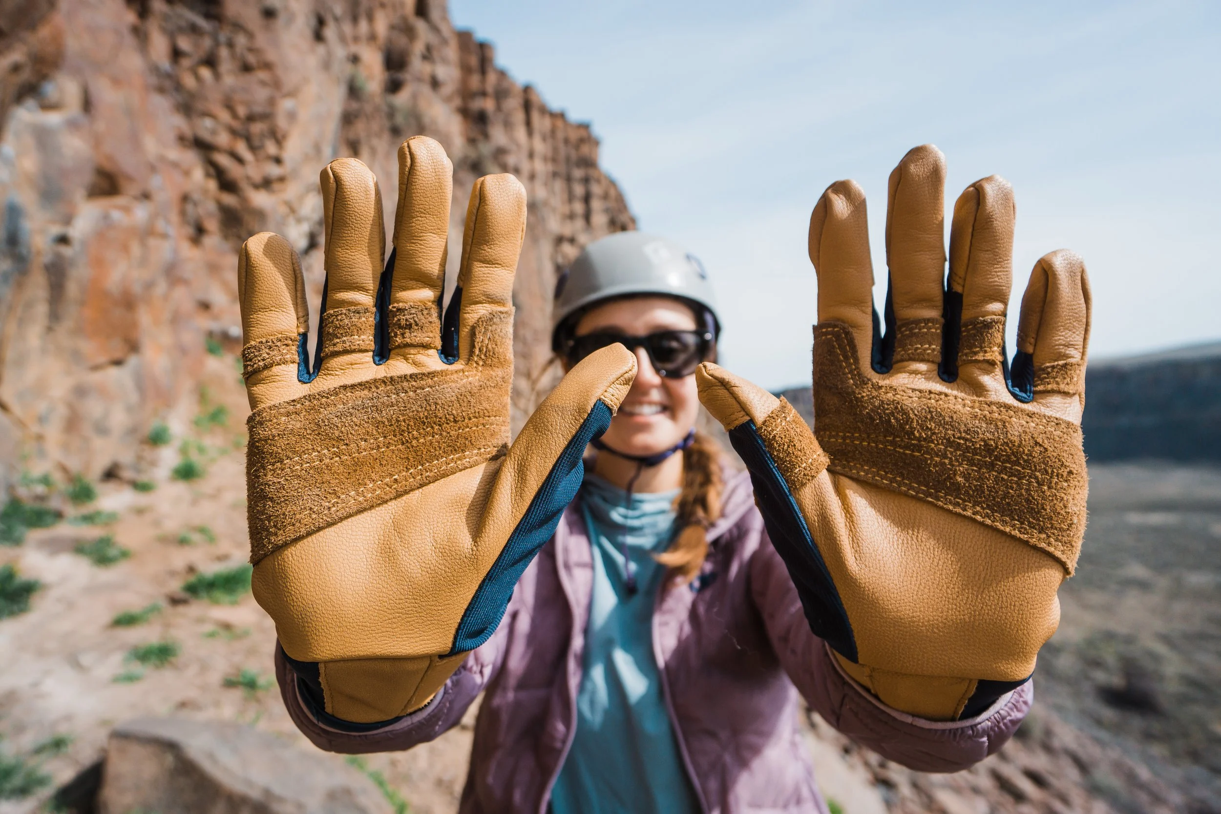 A woman wearing a helmet, sunglasses, and climbing gloves, holding her hands up with palms facing the camera, outdoors with rocky cliffs in the background.