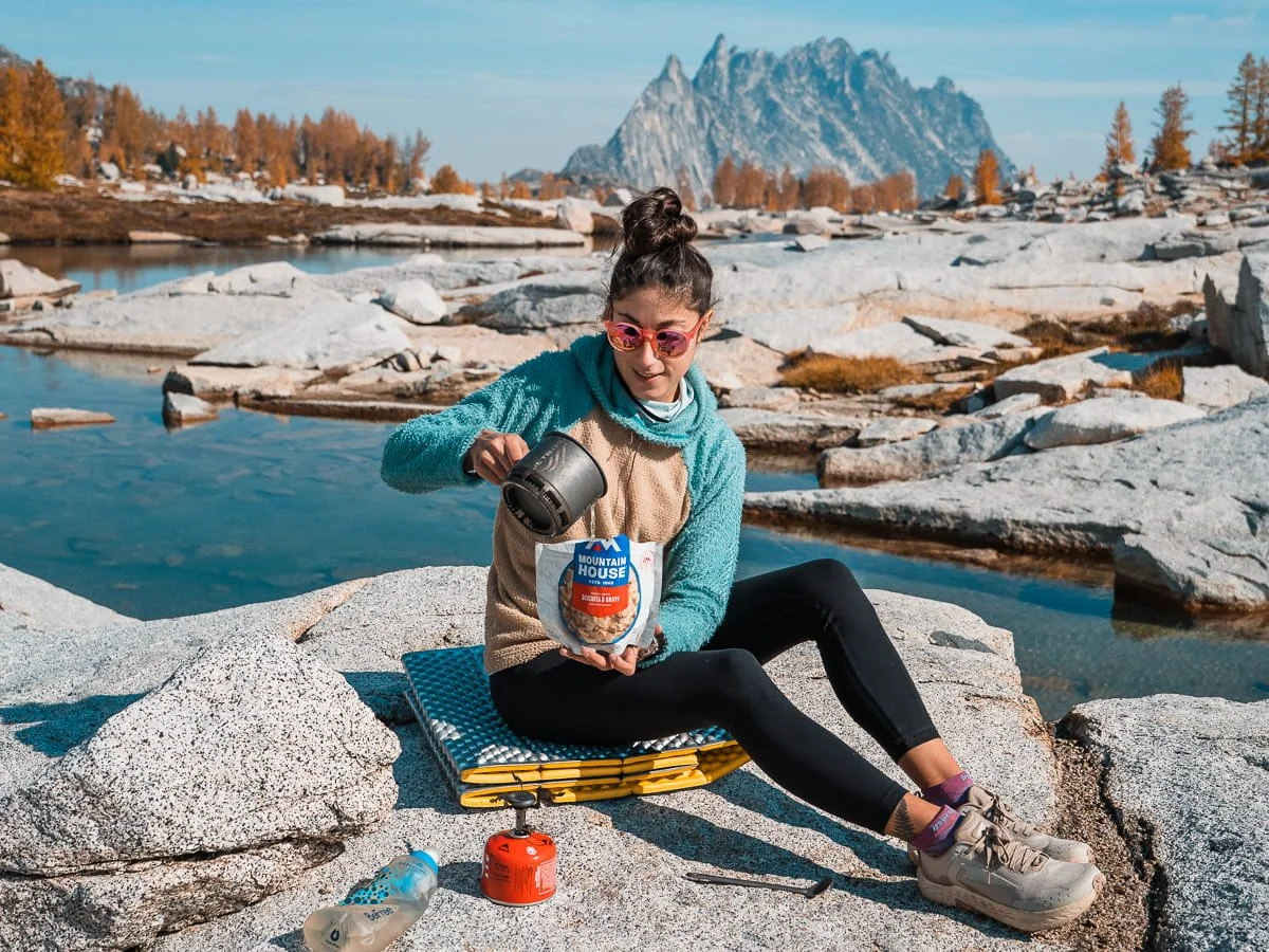 a woman making a freeze dried backpacking meal in the core enchantments with water and peaks surrounding her