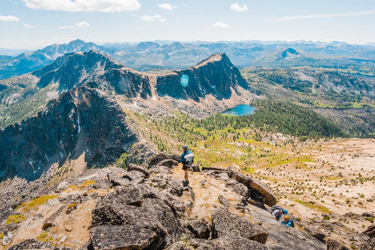 a backpacker looking out at a view of the pasayten wilderness from on cathedral peak. A long rocky ridge and an alpine lake in the distance.