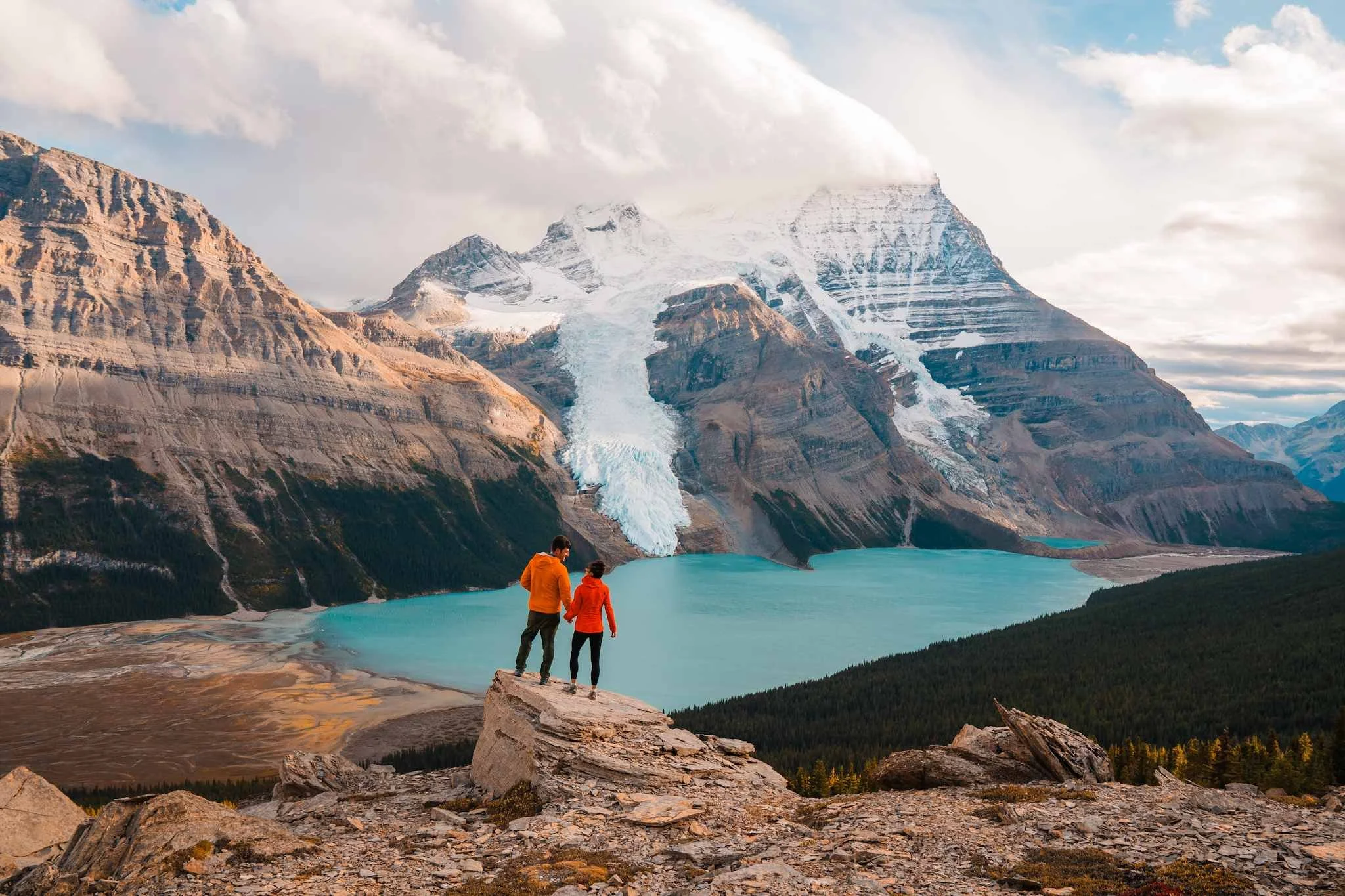 a couple in bright jackets, standing on a rocky view point overlooking a bright blue alpine lake and glaciated peak in the background
