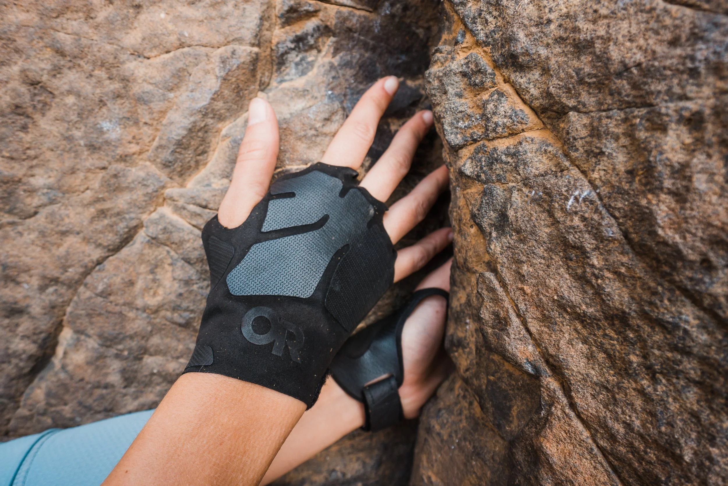 A person wearing a black and gray climbing glove is gripping a rough, brown rock face while rock climbing.