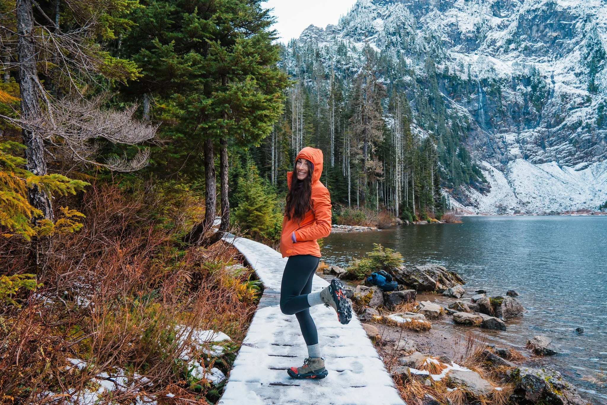a women on a snowy boardwalk next to a lake with trees and snow in the background. She is wearing a bright orange jacket with the hood on, black leggings, hiking boots, and microspikes