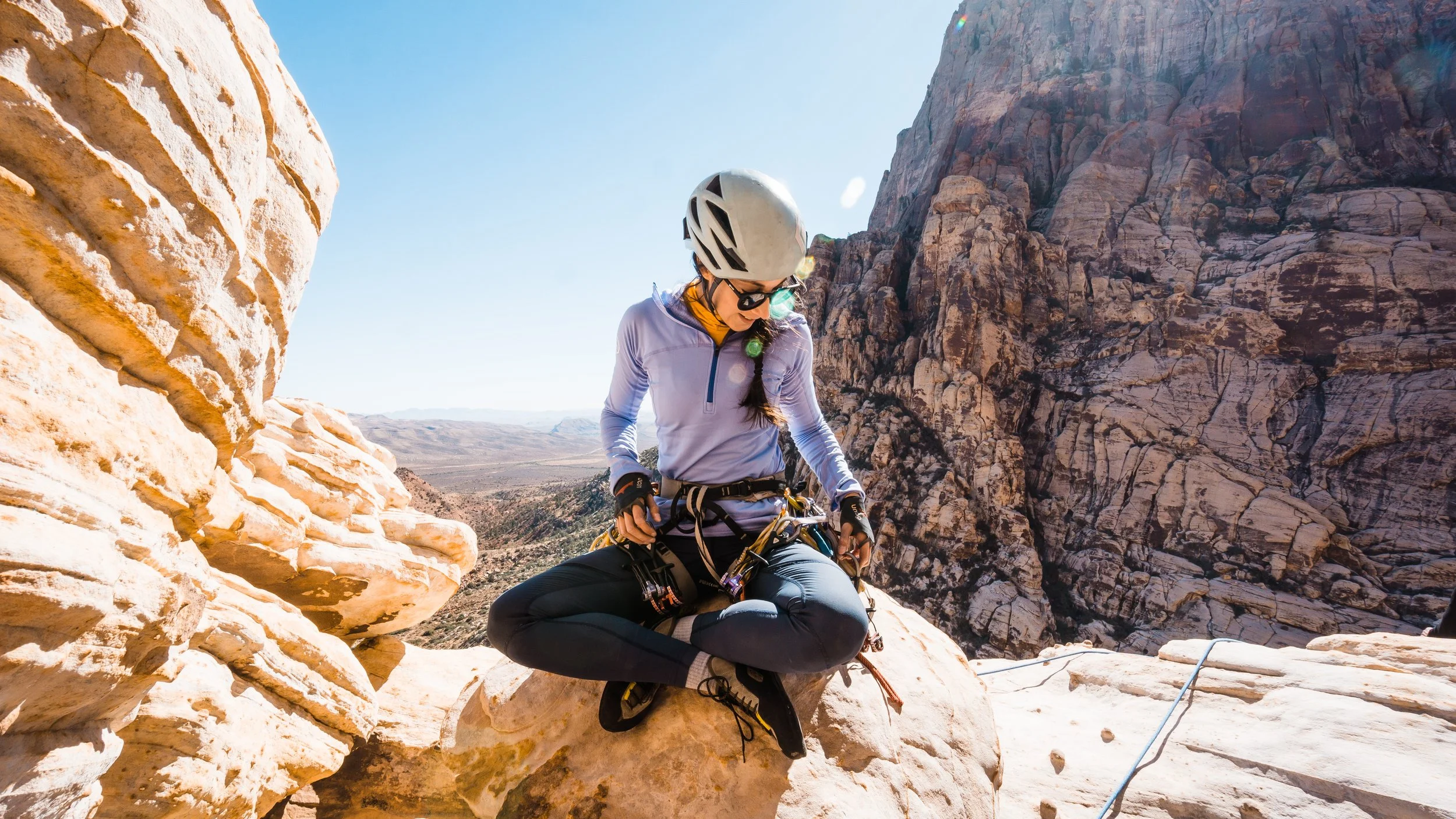 A woman wearing a helmet, sunglasses, and outdoor climbing gear, sitting on a rock in a desert canyon landscape with large rock formations and mountains in the background.