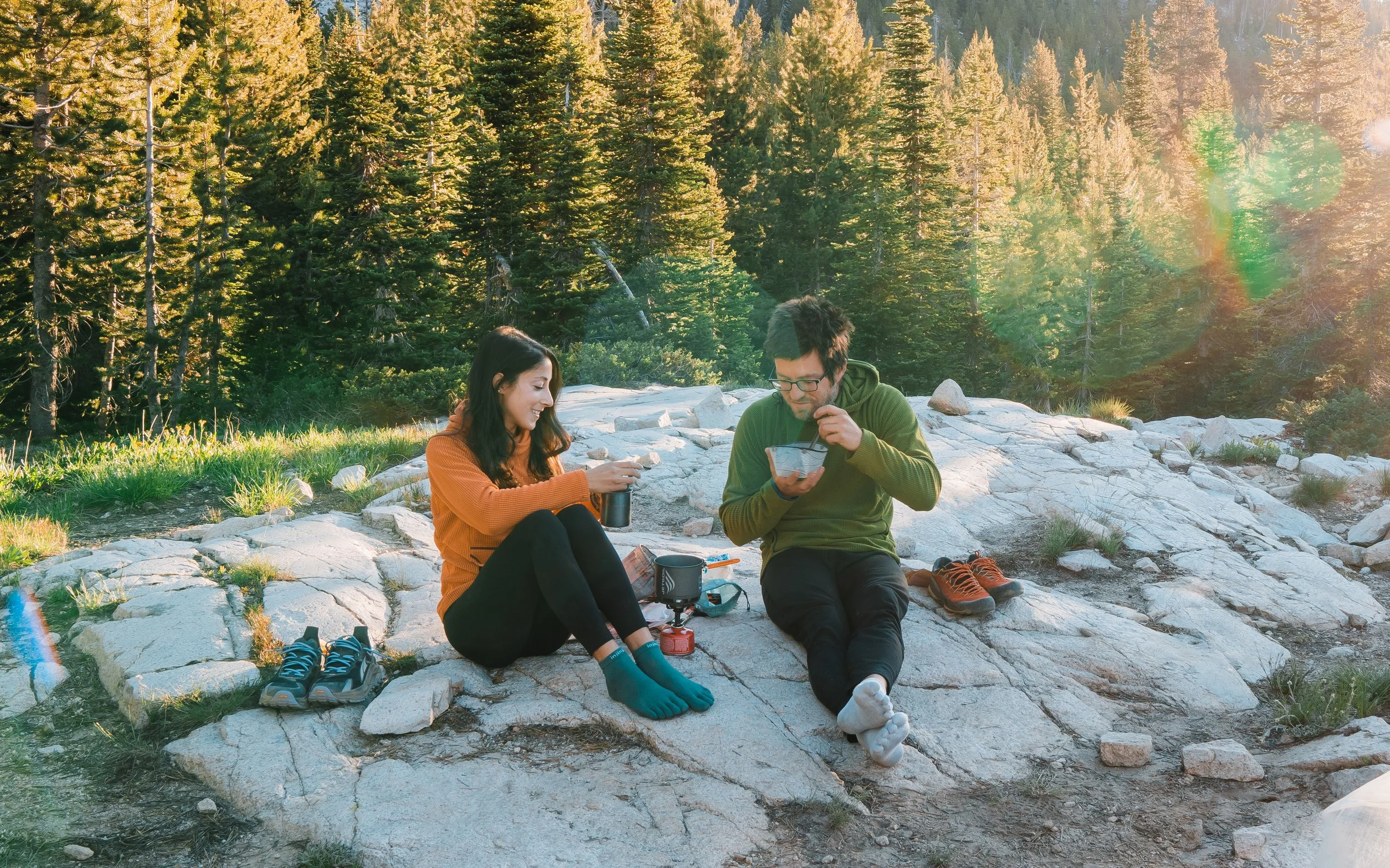 A couple sitting on rock slabs eating breakfast on a backpacking trip with pine trees in the background