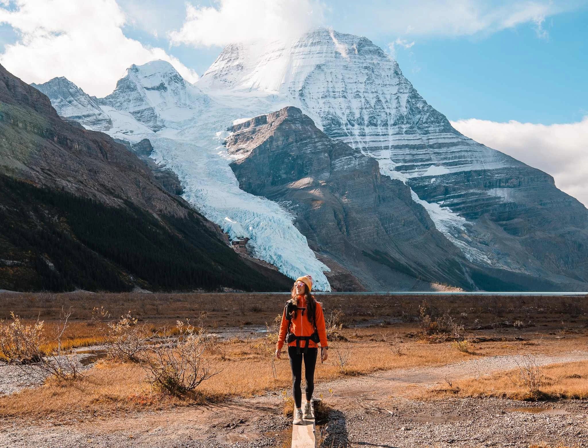 a women in bright hiking clothing on a small bridge in front of berg lake with mount robson in the background