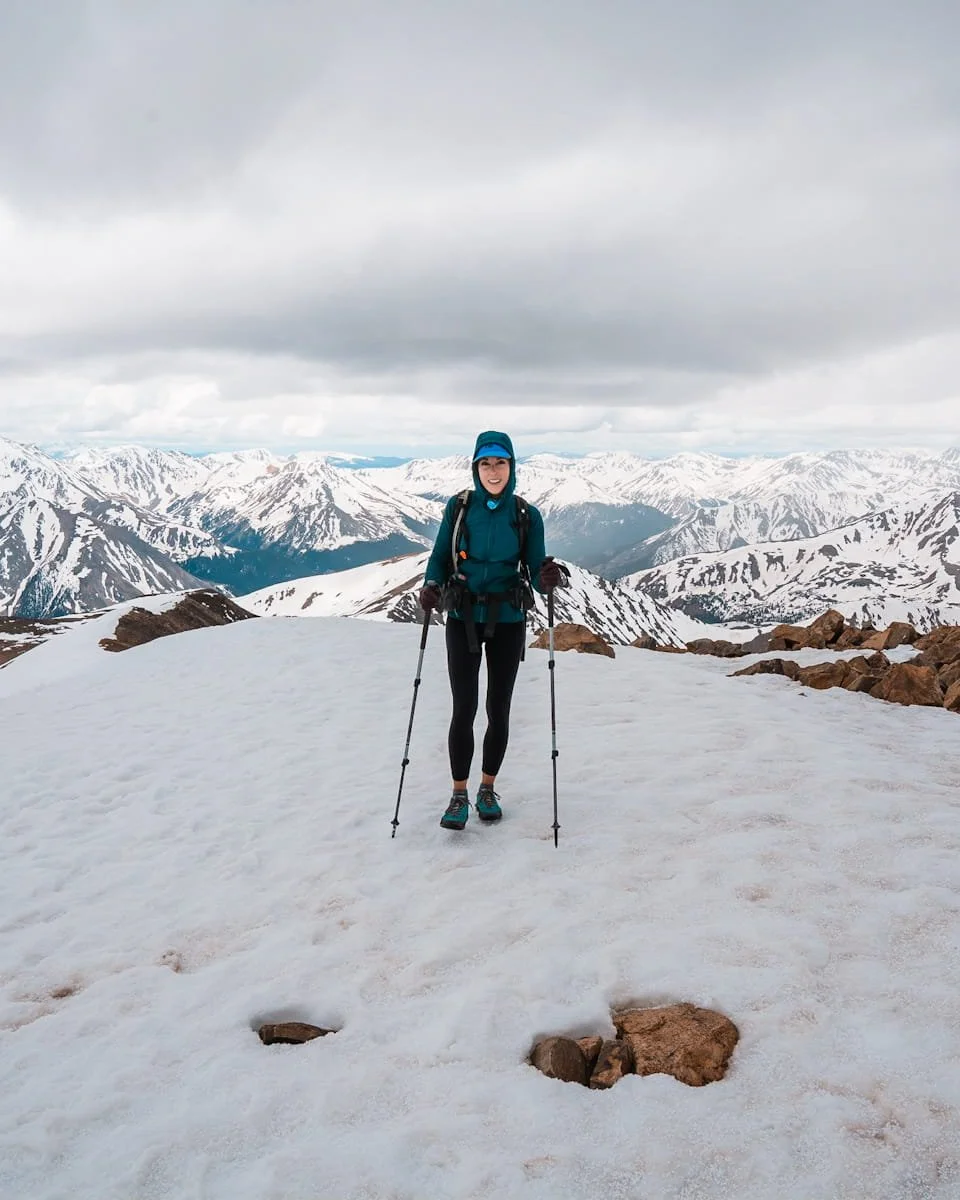 a woman all bundled up, hiking on snow at the summit of Mount Elbert with the colorado rockies in the background
