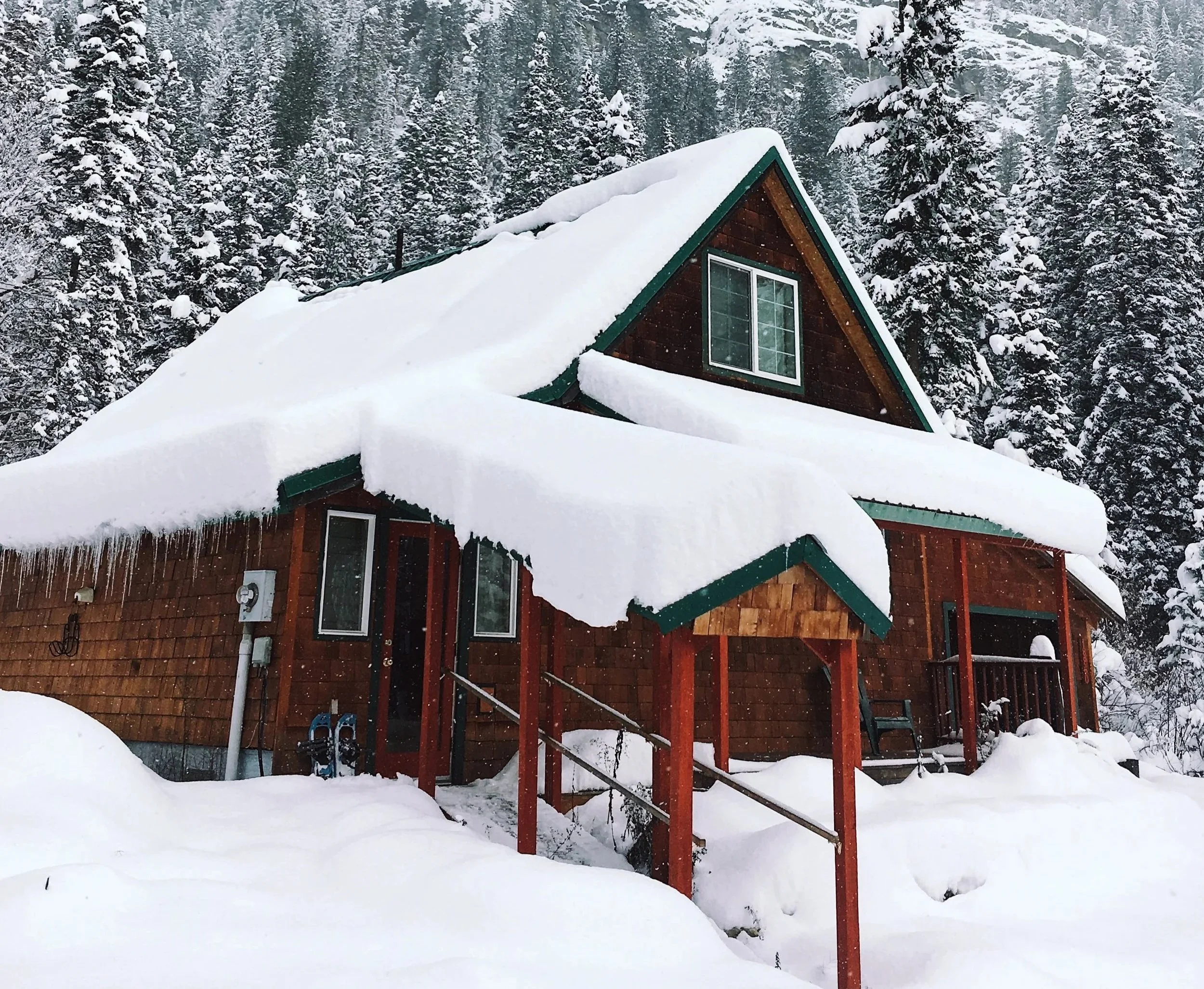 A cabin covered in snow with icicles coming off the roof and snowy trees in the background