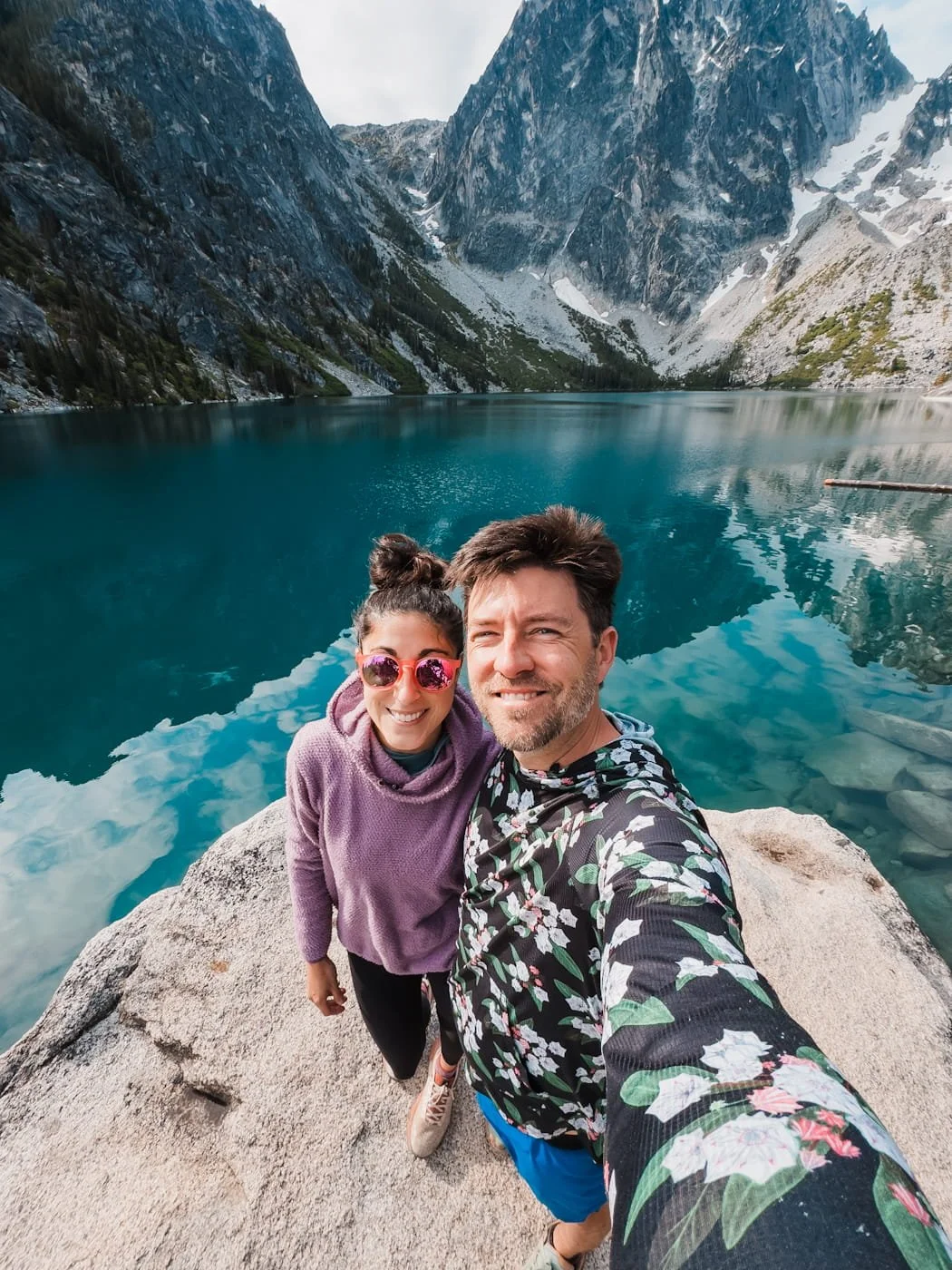 a couple standing in front of a blue alpine lake with granite peaks in the background