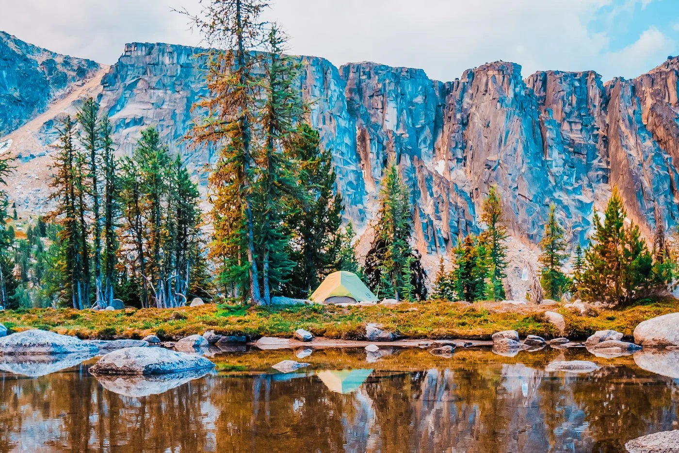 a tent reflecting in a tarn with pine trees and a rock face behind