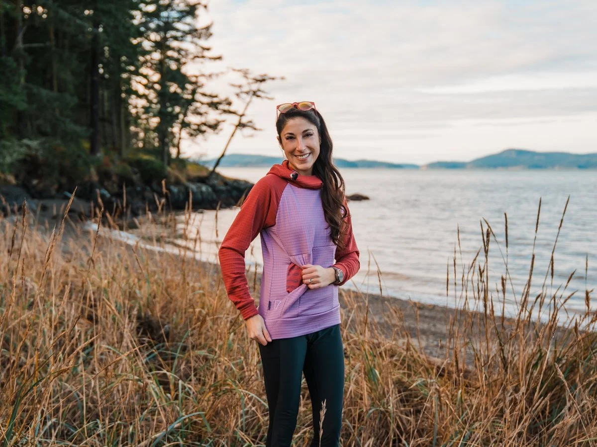 A woman wearing a color blocked grid fleece in light purple and dark pink, showing off the pocket, with a PNW coastal beach in the background