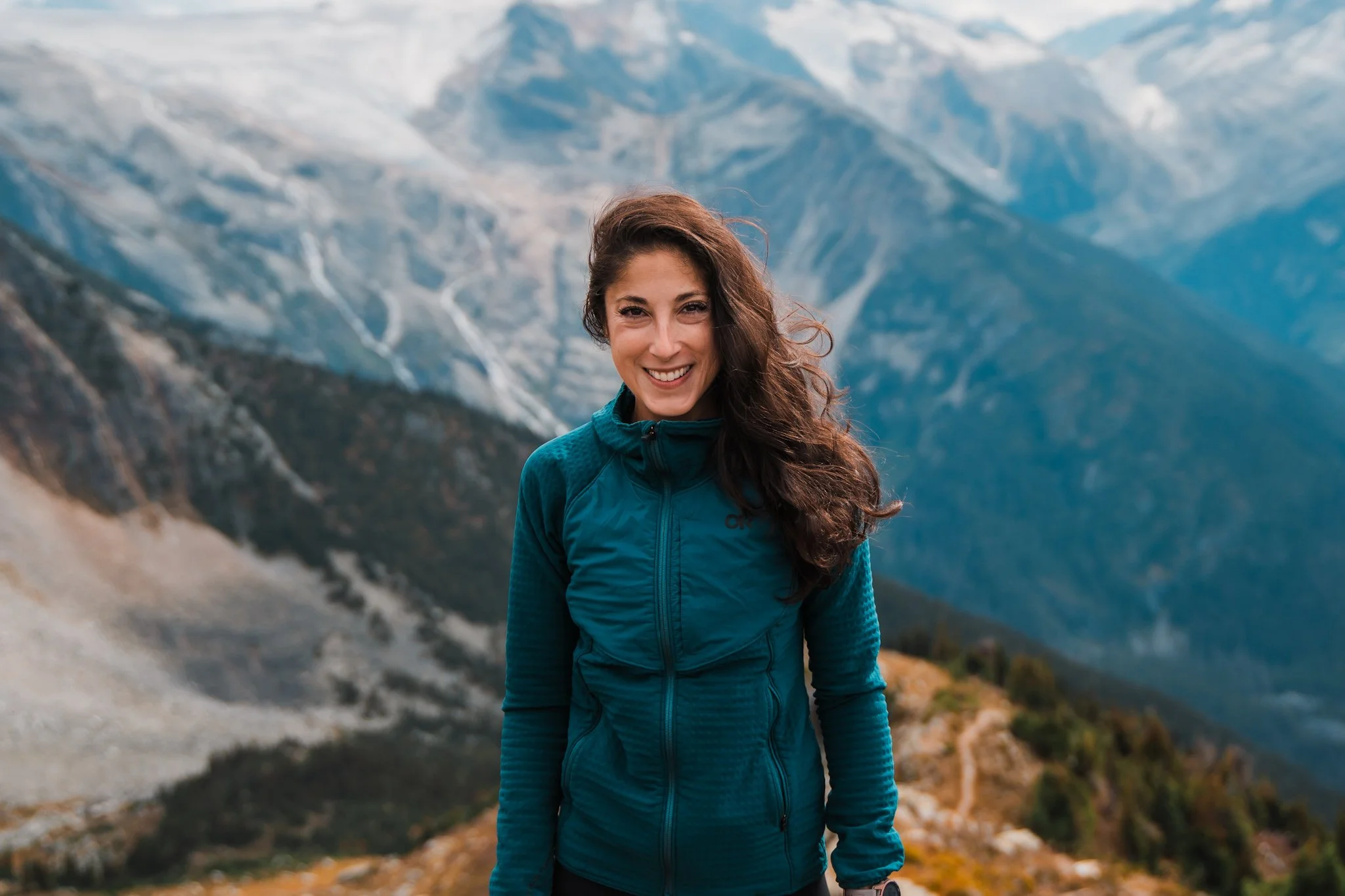 A women in a dark turquoise full zip fleece with brown hair blowing in the wind, standing in front of large mountains on a hiking trail in canada