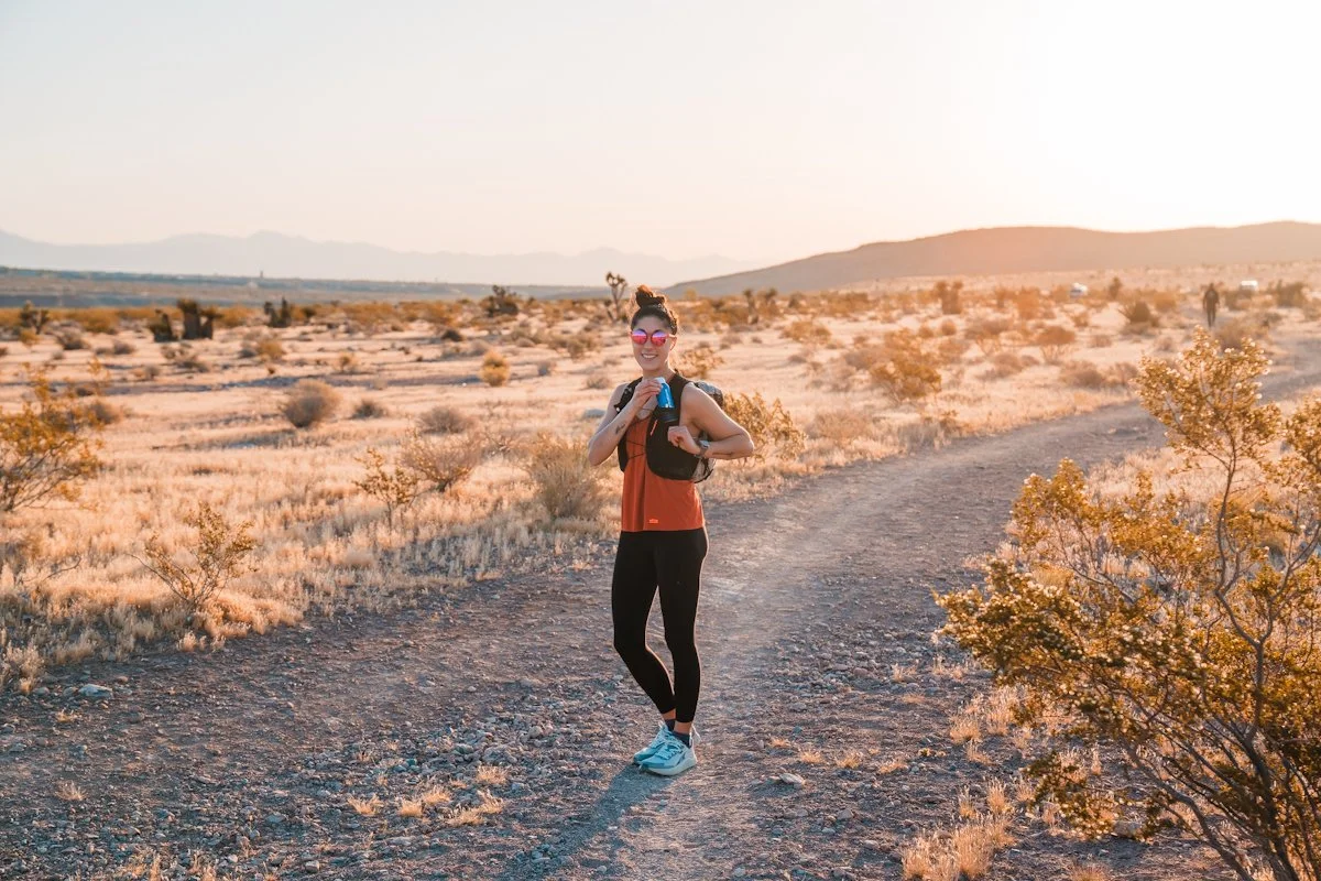A woman in athletic wear, wearing sunglasses and a backpack, standing on a dirt trail in a desert landscape during sunset, smiling and holding a drink.