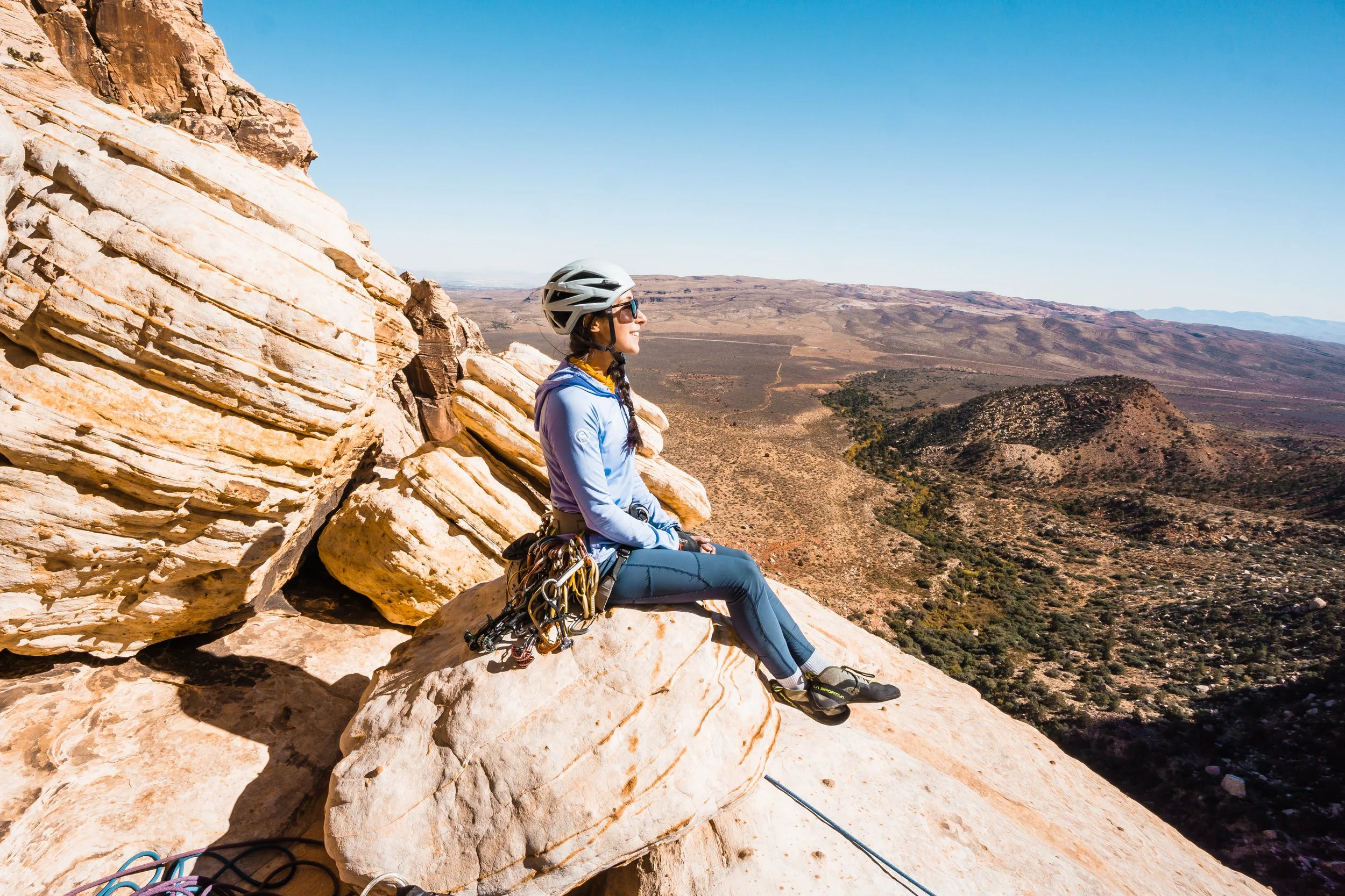 A female rock climber sitting on a large rock ledge on a mountain, wearing a helmet, sunglasses, and climbing gear, with scenic desert and mountain landscape in the background.