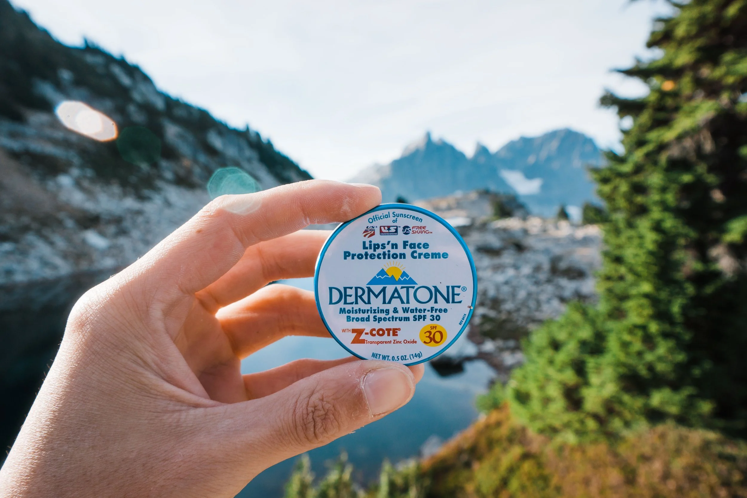 A hand holding a container of Dermatone face protection cream with a scenic mountain and lake in the background.