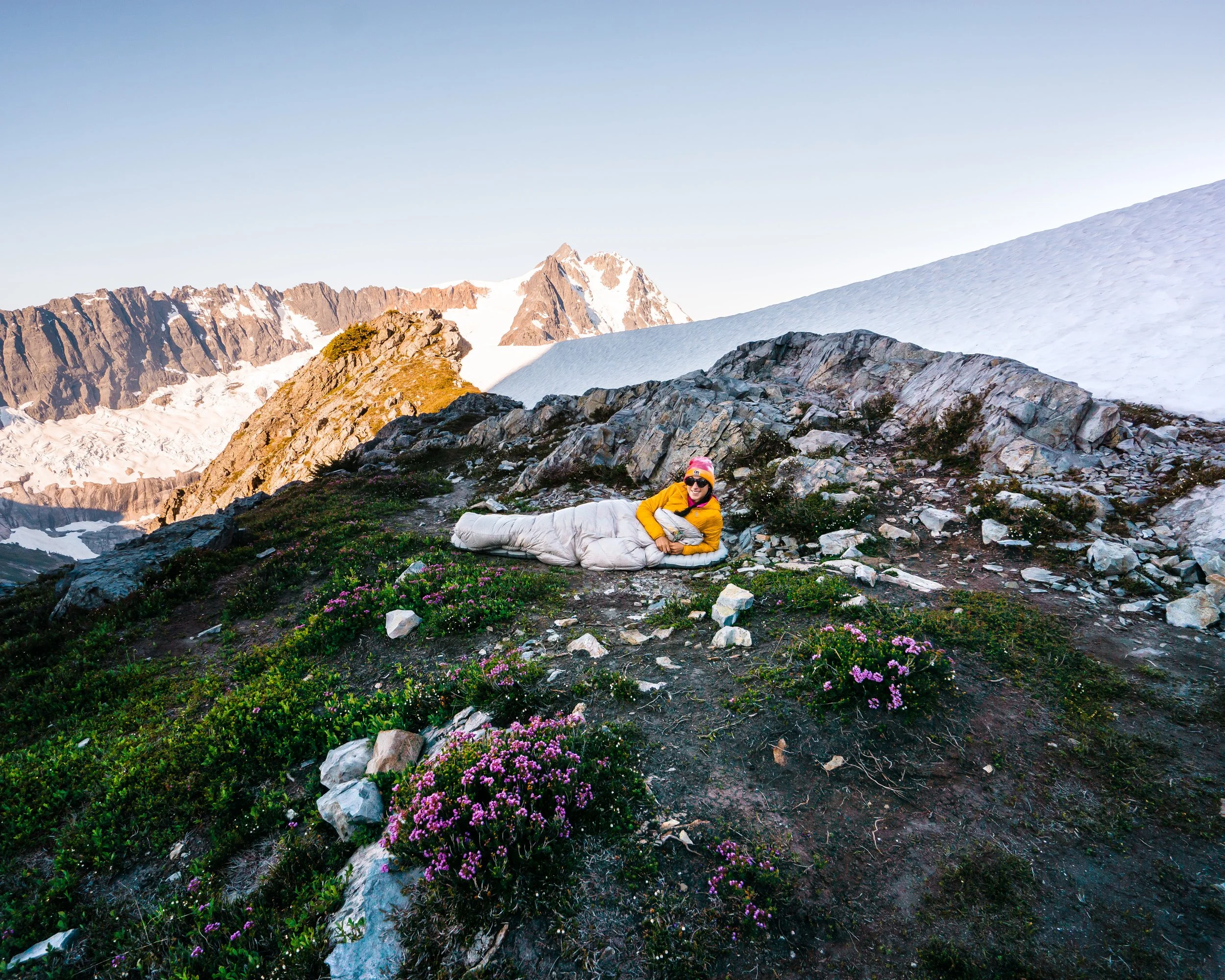 Person lying on the ground in a mountainous area with snow, rocky terrain, green plants, and purple flowers, wearing a yellow jacket, white sleeping bag, pink hat, and sunglasses.