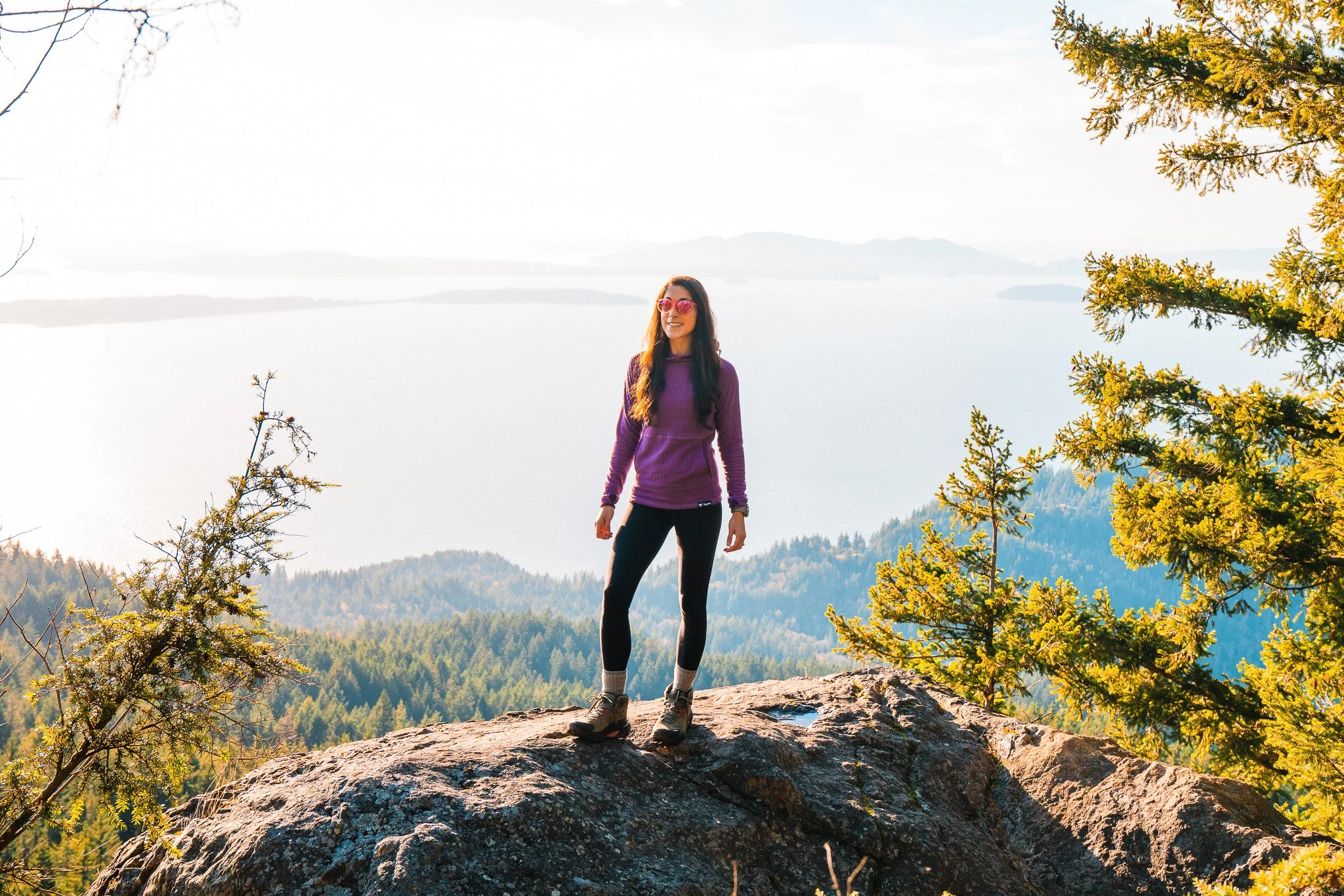 a woman standing on a rock cliff above the sound, wearing a purple grid fleece and black leggings