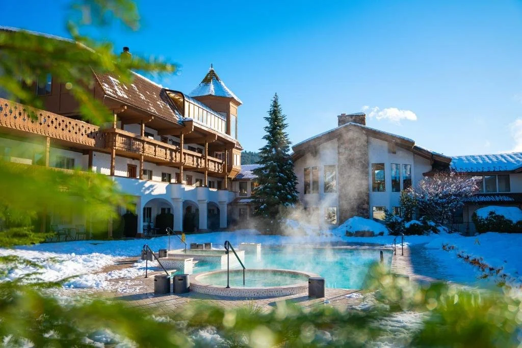 A hot tub and pool with steam coming off and snow around with the enzian hotel in the background