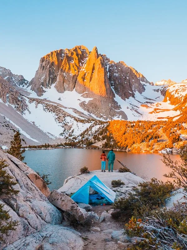 A blue tent and a couple standing and taking in the view of an alpine lake and mountain in the background