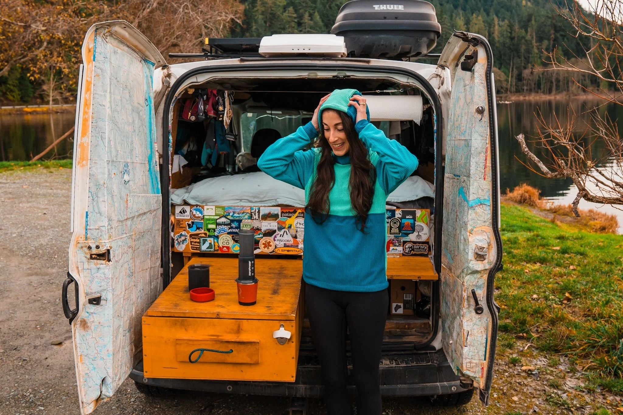 a woman standing in front of a small campervan, making an aeropress coffee, wearing a blue and turquoise, color blocked fleece hoodie